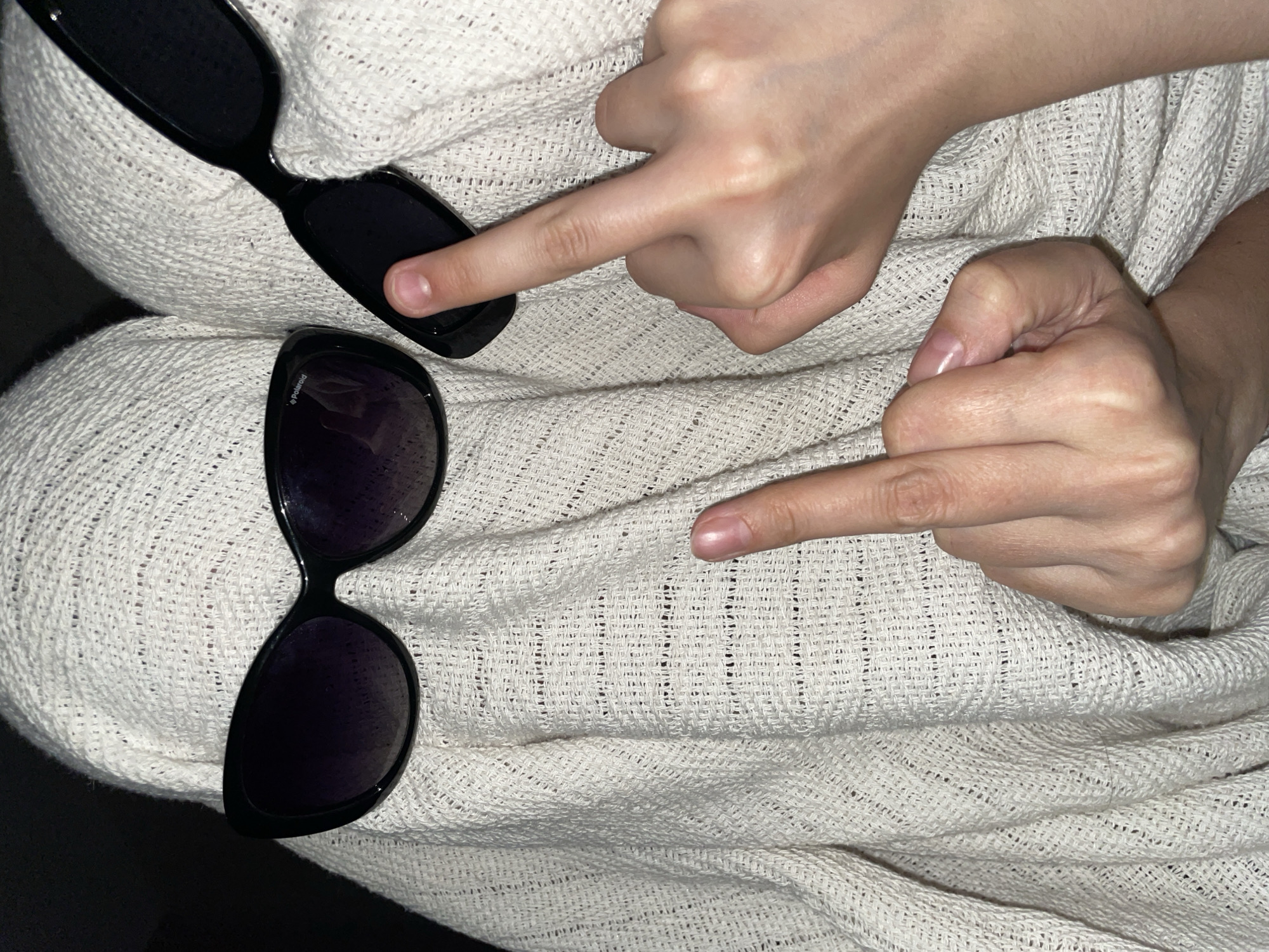 Two hands point to two pairs of black sunglasses lying on a light beige woven blanket.