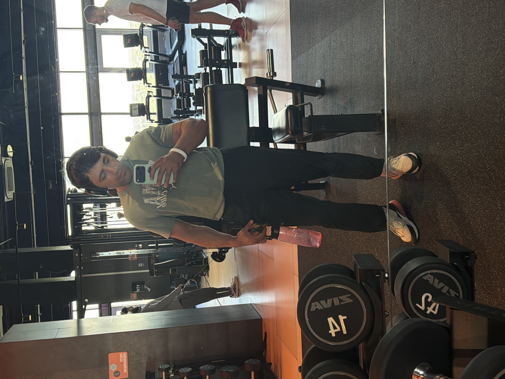 The image captures a mirror selfie taken inside a well-lit gym or fitness center in Krasnoyarsk, Russia, rotated 90 degrees clockwise. The primary subject is a man standing before a large mirror, holding a smartphone to take a picture. He wears a green t-shirt, black athletic pants, light-colored athletic shoes with orange-red accents, and headphones. A white wristband is visible on his right wrist, and he holds a translucent pink water bottle in his left hand.
The setting features large, multi-paned windows allowing bright natural light to fill the space, indicating daytime. The floor is covered with a dark, textured rubber material common in gyms. Various pieces of gym equipment, including weight machines and benches, are visible both directly and as reflections. In the foreground, a rack holds black dumbbells, some clearly marked with "AVIS," "4Г," and "SA."
In the background, reflected in the mirror, another man wearing a white t-shirt and dark shorts is visible, appearing to be near gym equipment. A third person is partially seen further back. An orange sign is visible in the background, with partially legible text including "ВНИМАНИЕ" (Attention) and "ВЫМОЙТЕ РУКИ" (Wash hands), suggesting hygiene instructions. The overall scene depicts a modern and functional gym environment.