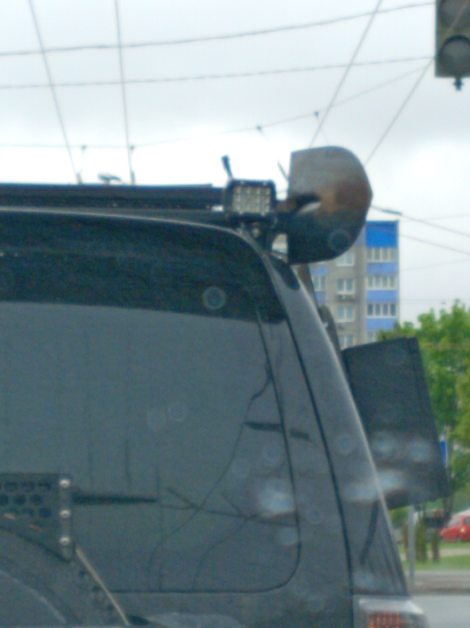 The image captures the back of a dark-colored SUV with various attachments on its roof and rear. A shovel and an LED light bar are mounted on the roof rack. On the rear, there's an angular accessory attached to the side of the SUV. The scene appears to be in an urban area, with a multi-story building visible in the background. Other vehicles, including a red car, are also present. The sky is overcast, suggesting possible rain, evident by droplets on the SUV's surface. Overhead wires are visible against the sky.
