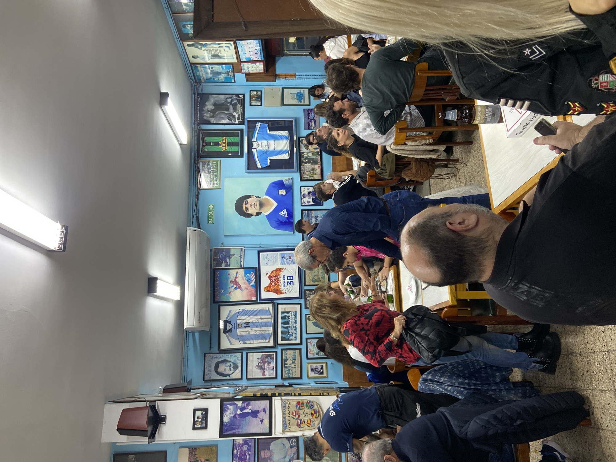 The interior of a restaurant or bar in Buenos Aires, Argentina, is filled with customers seated at tables. The walls are covered with framed images, posters, and memorabilia related to Diego Maradona and Argentinian soccer. Patrons are sitting closely together at tables, some are looking at their phones, others are interacting. A man in a black shirt stands near the front, and the top of a woman's blonde head is visible as well. There are fluorescent lights on the ceiling, and an air conditioning unit is mounted high on the wall. A sign indicates "Salida" (Exit). A bottle of Patagonia Lager del Sur is visible on a table, next to a paper with a telephone number printed on it.