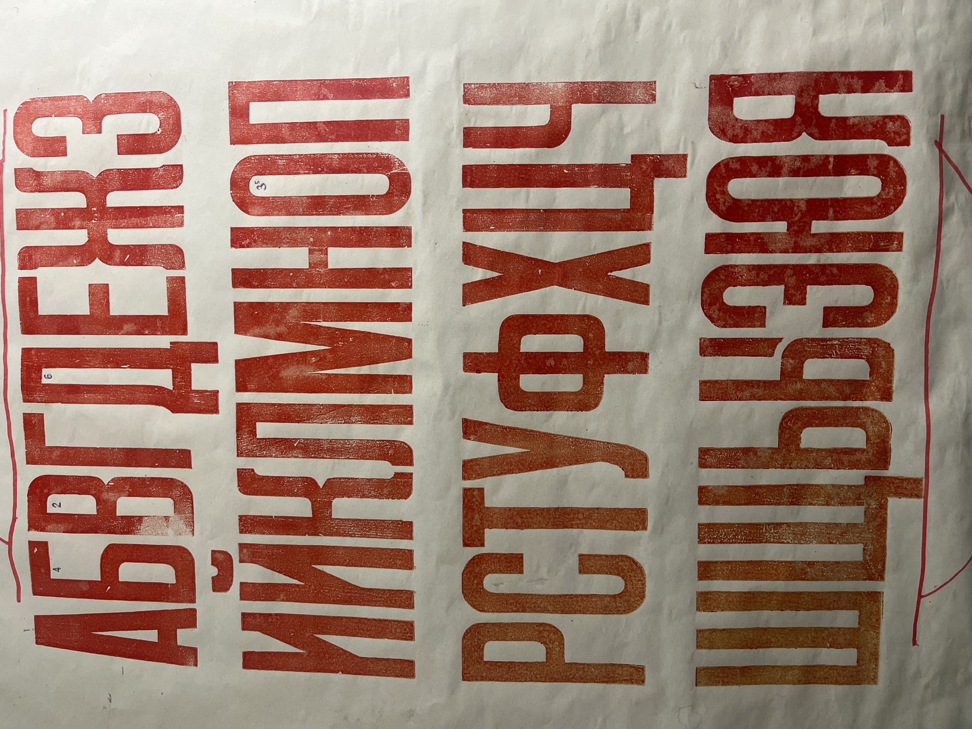 The image shows a large sheet of white paper with red, block-style lettering printed on it. The lettering appears to be in the Cyrillic alphabet and is arranged in four horizontal lines. Some of the letters have small numbers printed above or next to them, possibly indicating their order or a numbering system. The printing technique appears to be some form of relief printing, as evidenced by the textured and somewhat uneven ink coverage. There are no people, actions, or interactions visible in the image. The environment is a plain white background, suggesting this is a piece of artwork, a proof sheet, or educational material. There is no discernible connection to the city of Pionersky, Russia, as the image content is purely typographic.