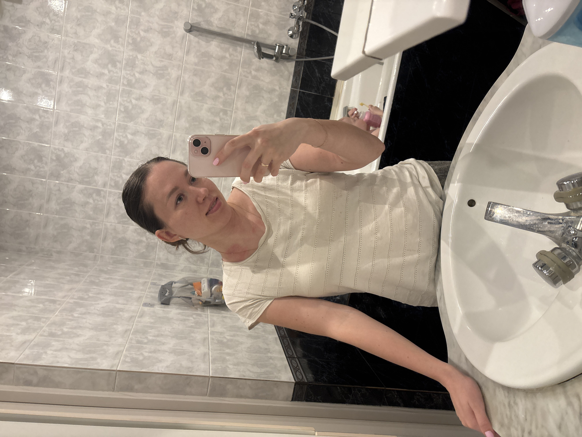The image depicts an adult female capturing a mirror selfie within a bathroom located in Ферма, Russia. The woman, appearing to be in her 20s or 30s, has dark hair pulled back and wears a light-colored, short-sleeved t-shirt with perforated horizontal lines. Visible red marks are present on her neck and collarbone. She holds a light pink smartphone with a dual-lens camera in her right hand to take the photograph; a light-colored ring is on her right ring finger and a thin bracelet on her right wrist. Her left hand rests on a light marble-patterned countertop beside a white ceramic sink with a chrome faucet.
The bathroom features light grey/white mottled square tiles on the upper walls, with a dark grey/black marble-patterned tile section below, separated by a decorative border. A white bathtub with chrome shower fixtures is visible in the background reflection, along with a pink plastic bottle inside the tub. A shark-shaped toothbrush holder containing an orange toothbrush is mounted on the tiled wall behind the woman's left side. A partially visible light blue soap dispenser is on the countertop to the right.