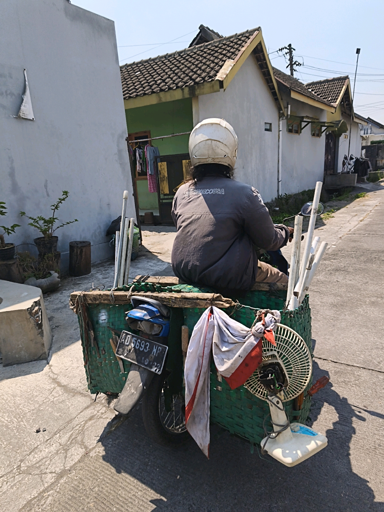 In Desa Sawahan, Indonesia, a person wearing a white helmet and a grey jacket is riding a motorbike. The motorbike is modified with a large green woven basket attached to the rear, carrying various items including several white pipes, a white electric fan, and some fabric. A blue seat and a license plate with the registration "AD 5693 NP" are visible on the motorbike. The background shows typical residential buildings with tiled roofs and white walls, a paved road, and a clear sky, suggesting a sunny day. The scene captures a moment of transportation and commerce in a rural Indonesian setting.
