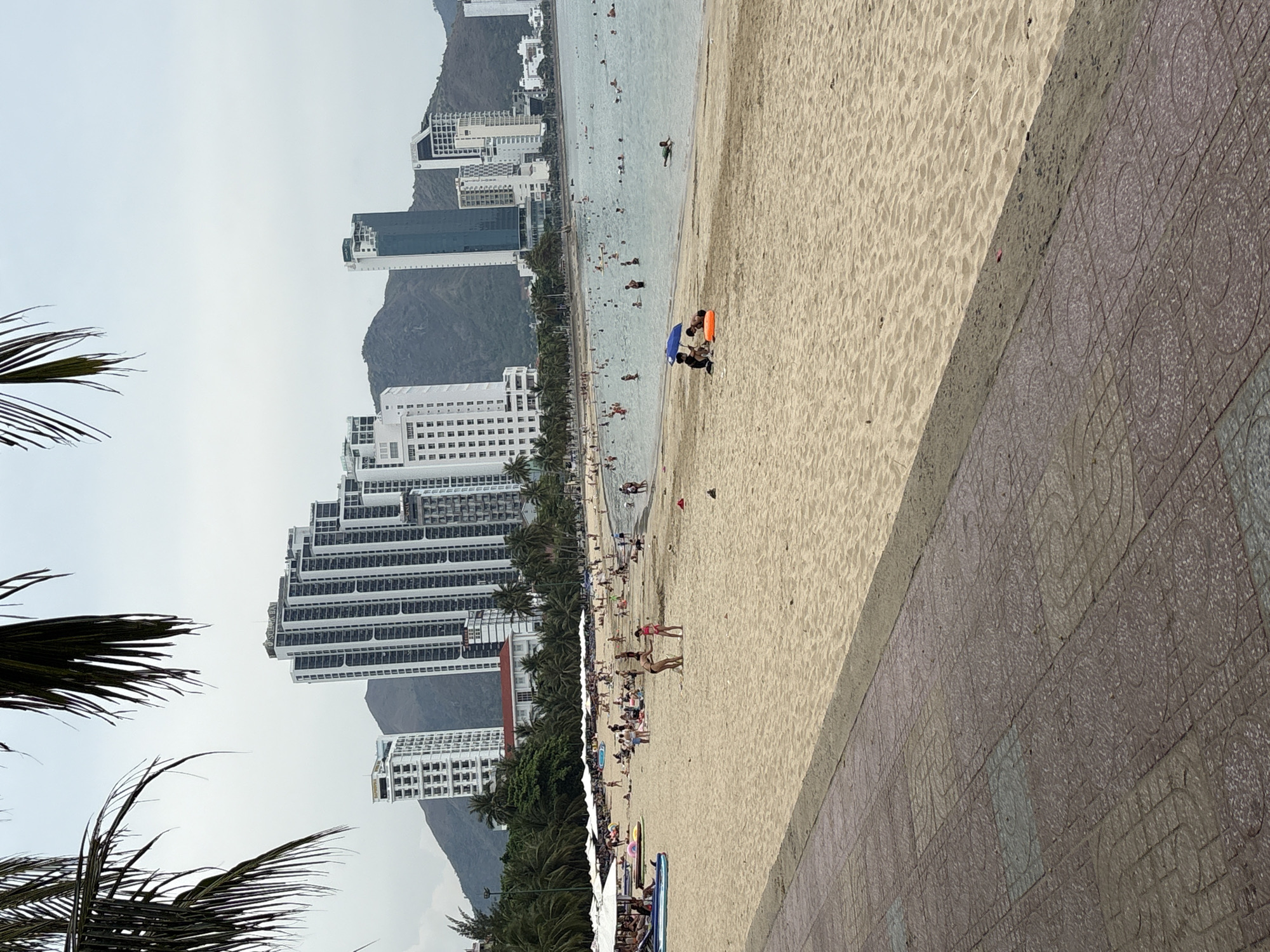 The scene is a beach in Bac Nha Trang Ward, Vietnam. In the background are buildings and a mountain under an overcast sky. A sandy beach stretches across the midground, populated with people swimming, walking, and relaxing. A stone walkway runs along the edge of the beach. Palm tree leaves are visible on the left.