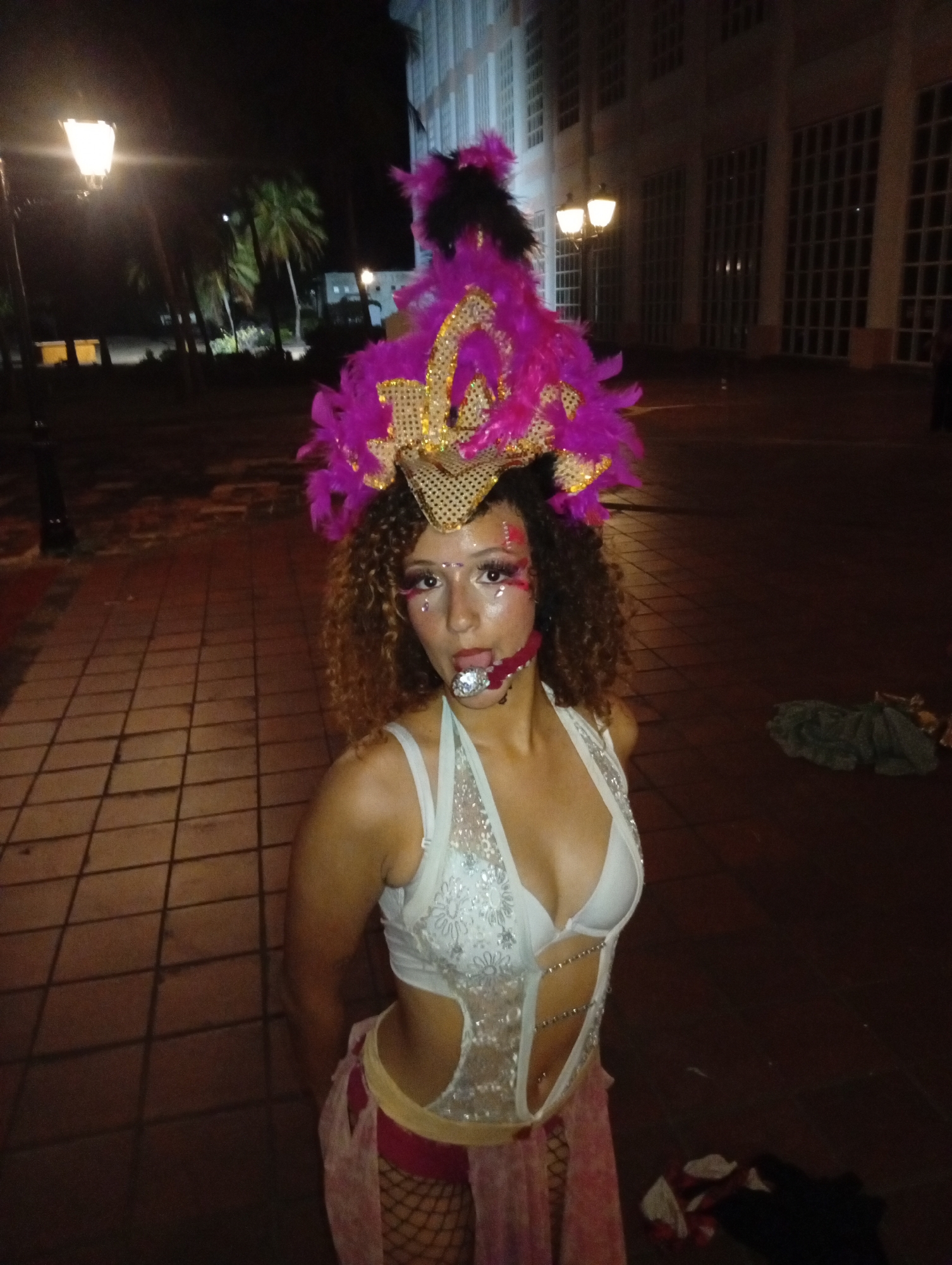 A person in costume stands in a public space in the city of Pedro González, Venezuela. The location is outdoors, at night, on a brick-paved area illuminated by lampposts. The person is wearing a white, sequined, revealing costume with a bra-style top, a pink wrap, and fishnet stockings. They have a large headdress with pink and black feathers and gold accents. Their face is decorated with glitter and a silver object in their mouth. Behind the person is a building with large windows. In the background, there are trees and additional lampposts. There are items on the ground to the right of the person.