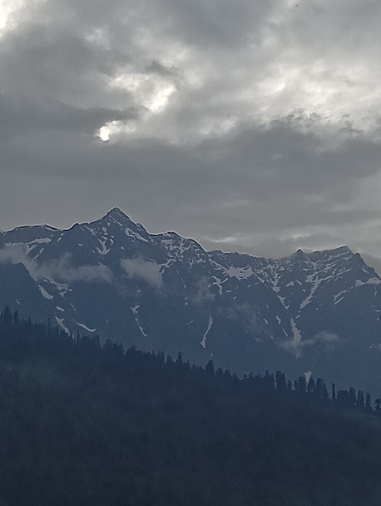 An overcast sky looms above a rugged, snow-capped mountain range. The clouds are a mix of dark grey and lighter patches, with a hint of sunlight breaking through in the upper left quadrant. The mountains themselves are imposing, with sharp peaks and jagged ridges. Streaks of white snow are visible against the dark rock, particularly on the higher slopes. In the foreground, a dense forest of dark green trees covers the lower slopes of the mountains, creating a stark contrast with the rocky peaks above. The overall impression is one of natural grandeur and a slightly dramatic, moody atmosphere. The location is specified as Vashisht, India.