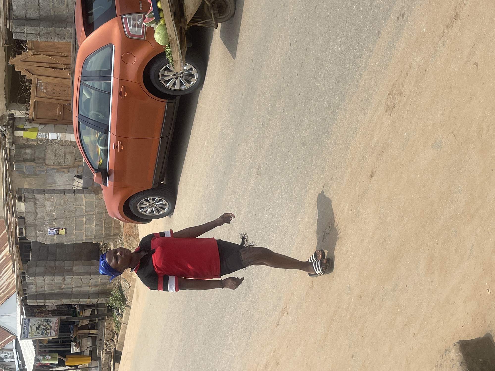 In a medium shot, a woman walks down a paved road in Abuja, Nigeria. She wears a blue headscarf, a red shirt with black and white stripes on the sleeves, and a short black skirt with frayed edges. She has on black and white striped sandals. The woman is walking on the right side of the road, facing left. Behind her, an orange SUV is parked on the right side of the road. A vendor's cart with fruits, including watermelons and some citrus fruits, is positioned next to the SUV. The background shows a row of buildings with corrugated iron roofs and concrete block construction. The road surface is asphalt in some parts and dirt in others.