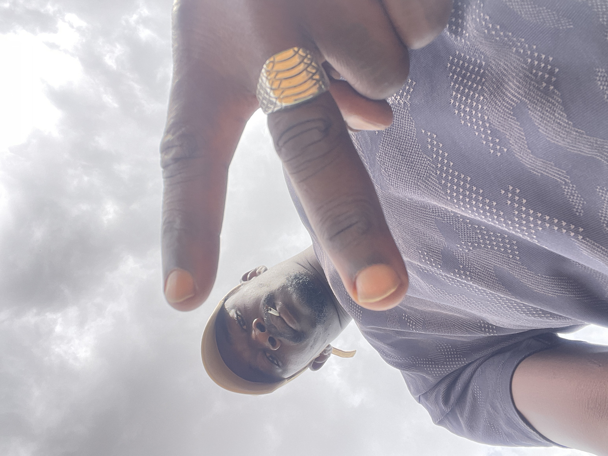 A low-angle shot depicts a person looking down at the camera. The person is dark-skinned and has a cigarette in their mouth and is wearing a tan cap. They are wearing a blue shirt with a patterned design and a gold ring on their finger. The background is a cloudy sky. The city of Durimi, Nigeria is not discernible from the image.