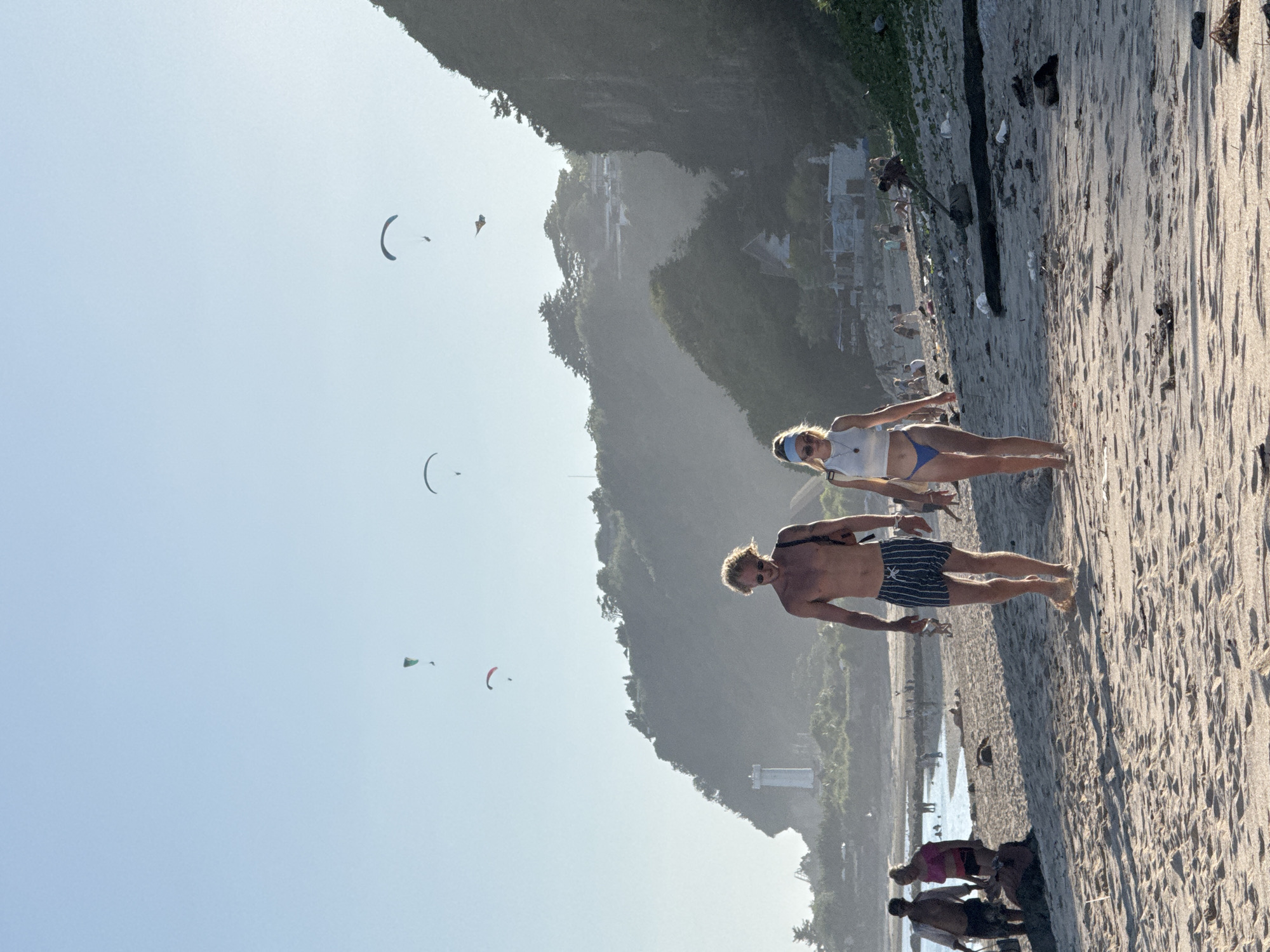 The image depicts a beach scene in Kuta Selatan, Indonesia. Two people, a man and a woman, are walking on the sandy beach. The man is shirtless, wearing striped swimming trunks, and is fair-skinned. The woman has blonde hair and wears a white top and blue bikini bottoms. Paragliders are visible in the sky above the beach. The background features a hillside covered in vegetation and some structures. Other people are visible on the beach near the water.