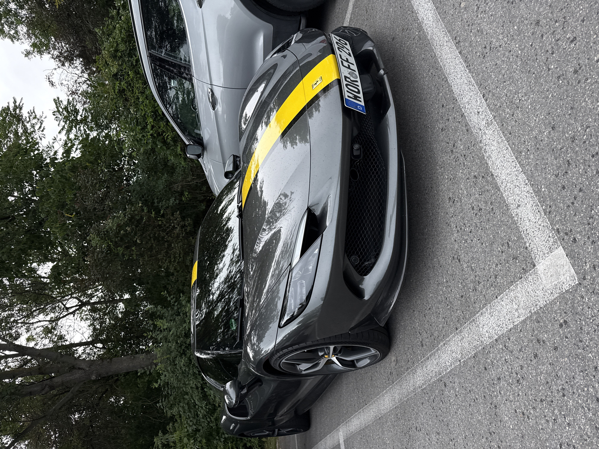 A dark grey Ferrari sports car with a yellow stripe down the middle of its hood is parked in a lot. The car is facing the camera and is partially obscured on the right by a silver SUV. The Ferrari has its headlights on and a license plate that reads "WOR FF 296". The parking lot appears to be paved with asphalt and has white parking lines. In the background, there are trees and foliage. The sky is overcast.