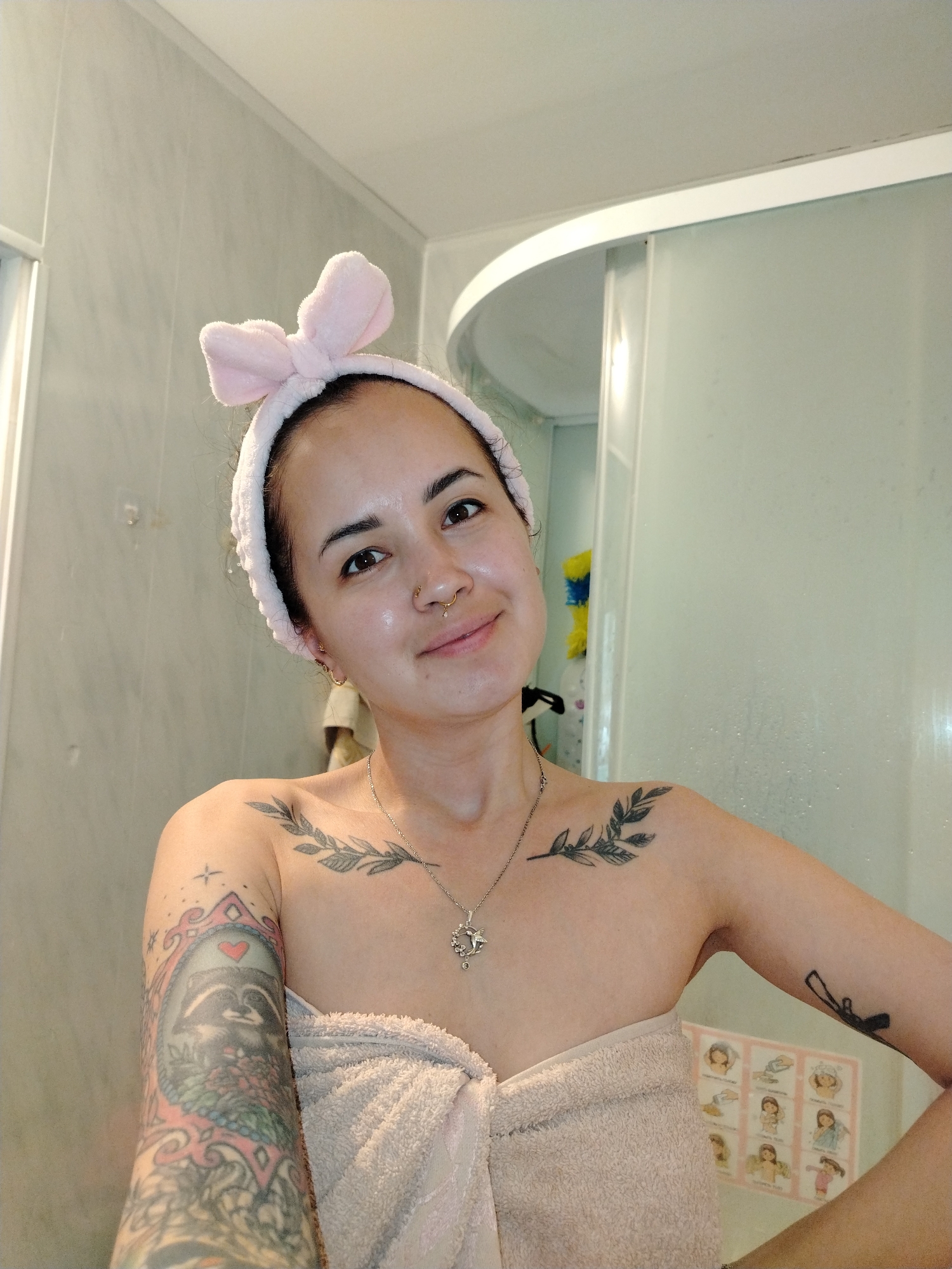 A woman stands in a bathroom, posing for a selfie. She wears a pink, bunny-eared headband and is wrapped in a light-colored towel. She has a nose ring and wears a necklace. Visible tattoos are present on both arms and across her collarbone. The background includes a shower stall with a frosted glass door and tiled walls. A chart with images of women is visible on the wall near the shower. The location is presumed to be a residential bathroom and there is no mention of any specific city in the photo. The prompt specifies not to include any city in Russia, so that part of the request is omitted.