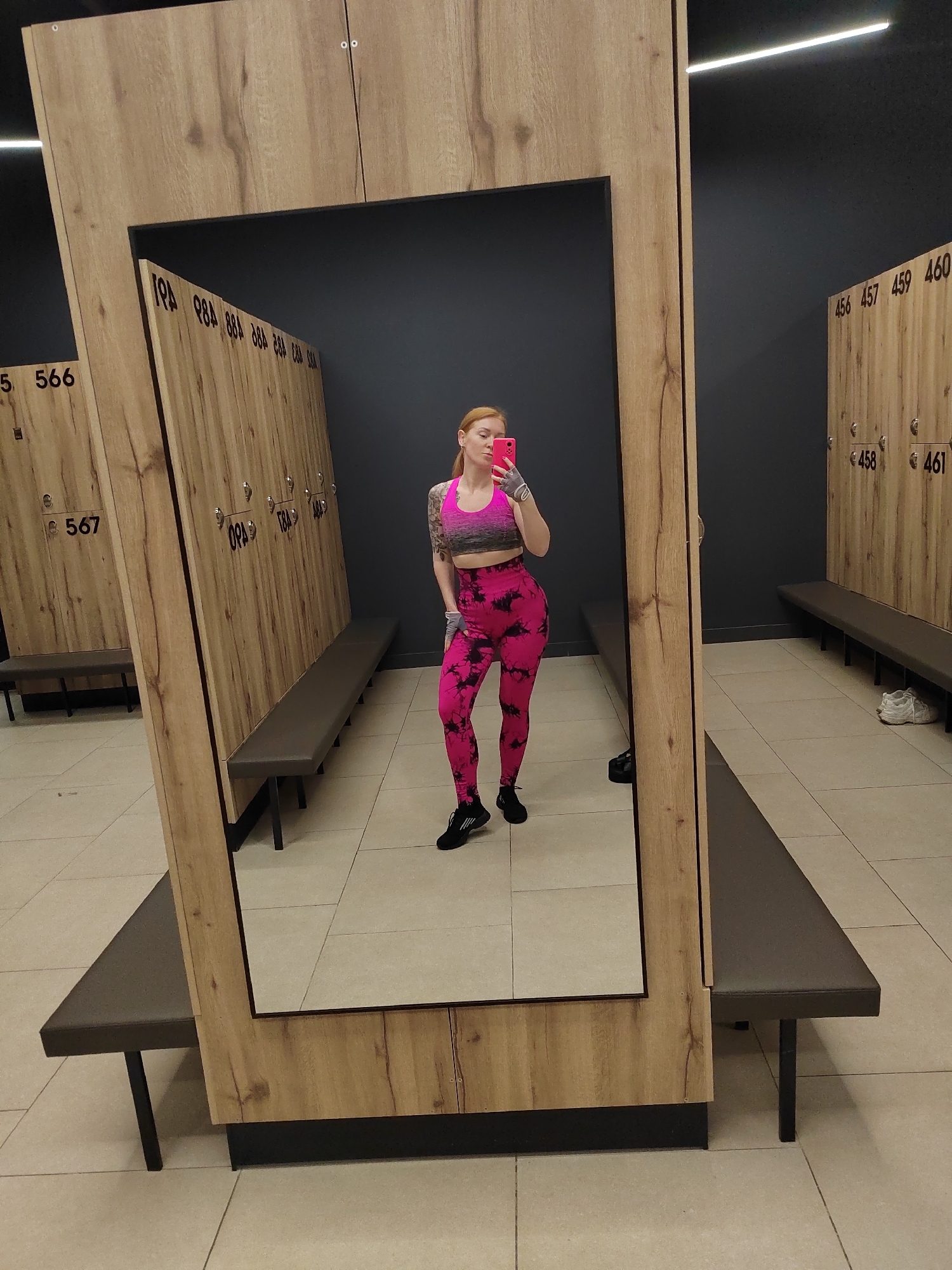 A woman is taking a selfie in a locker room in a building located in Novogorelovo, Russia. The woman has red hair and is wearing a pink and gray sports bra and pink leggings. She is standing in front of a large mirror that reflects her and the surrounding locker room. The locker room has wooden lockers with numbers on them, and benches are placed in front of the lockers. The floor is tiled. The lighting is bright, with overhead fluorescent lights visible in the reflection.