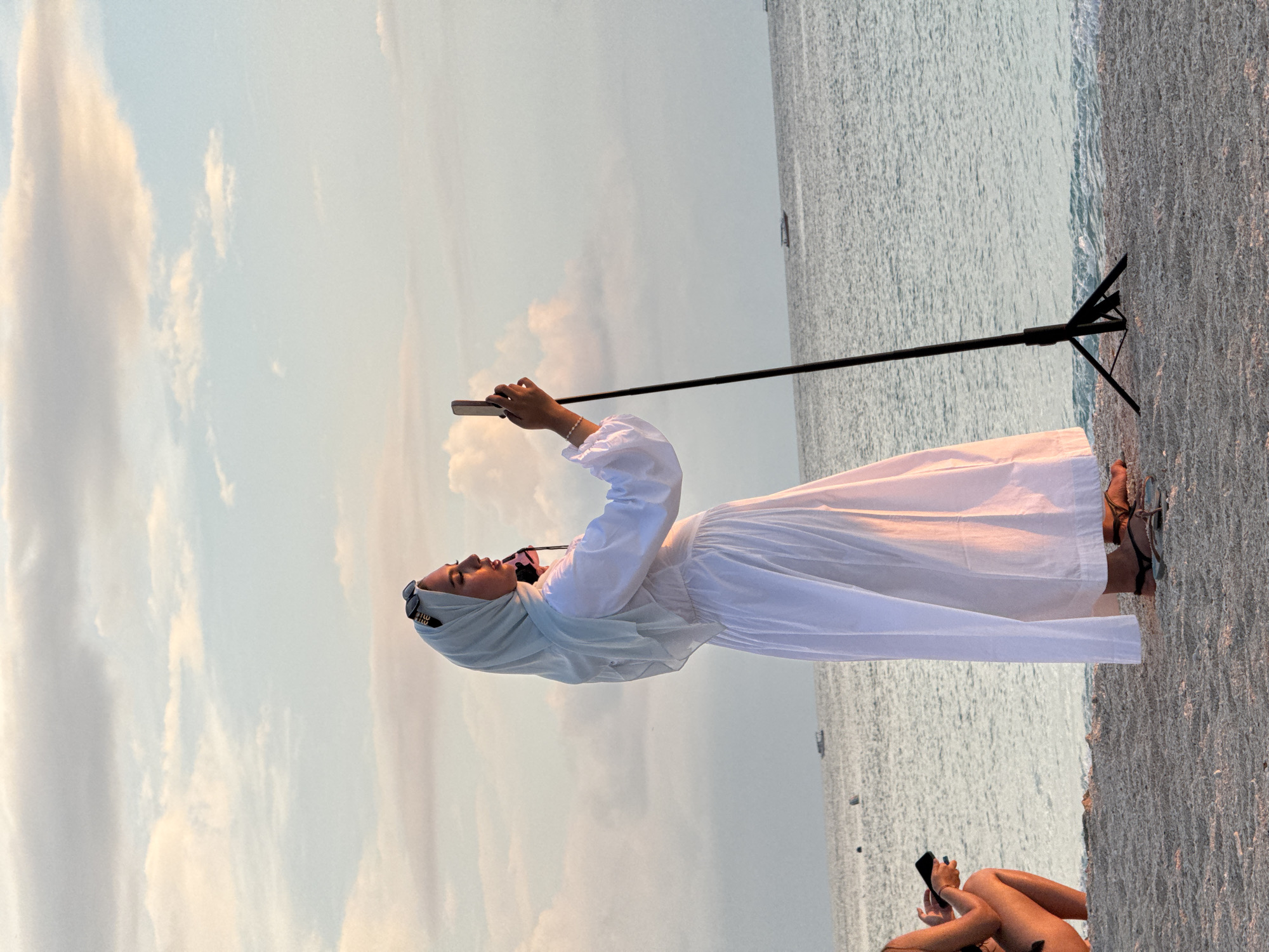 A person in a white dress and hijab is holding up a mobile phone on a selfie stick on a beach. Another person is seated on the beach in the foreground. In the background is water and cloudy skies. The location is in the city of Unknown, Indonesia.