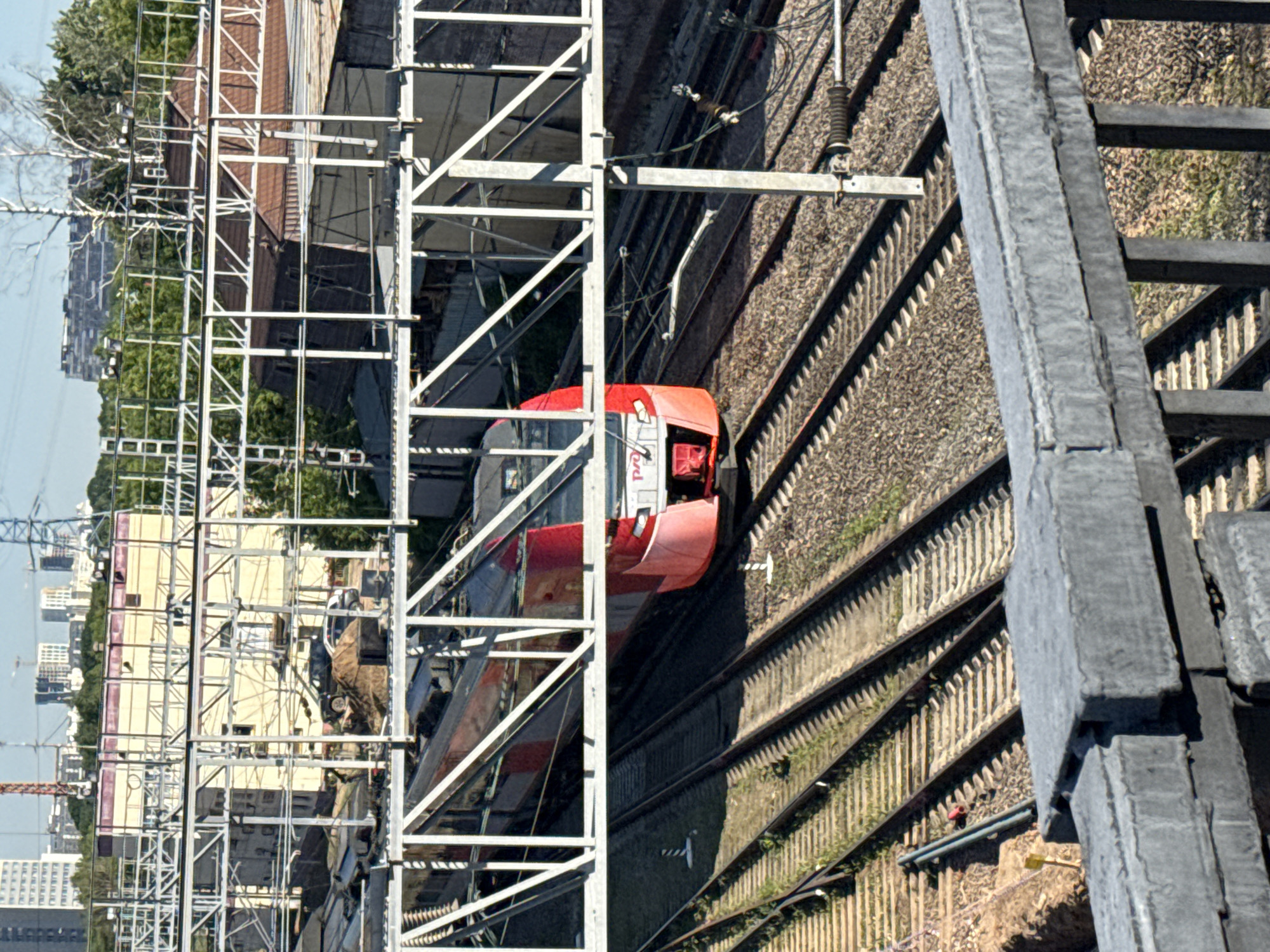 This image provides a high-angle, overhead view of a railway track and a train within an urban or industrial landscape, likely Moscow, Russia. The scene is bathed in bright daylight under a clear sky, evidenced by the distinct shadows cast by various structures.
A red and white passenger train, identifiable as an "Aeroexpress" service by the visible branding on its side, is positioned on the tracks. The front and side of one carriage are visible, suggesting the train is either stationary or moving slowly from left to right across the frame. Several sets of railway tracks, complete with ballast and concrete sleepers, run diagonally through the scene.
Overhead electrification lines (catenary system) are supported by a complex network of tall, gray metal gantries and poles that crisscross above and alongside the tracks. In the immediate foreground, a heavy, dark gray metal structure, possibly part of a bridge or an elevated walkway, partially obscures the tracks, featuring thick beams and steps.
The background on the left side of the tracks reveals a mix of buildings: some appear to be industrial or commercial with yellow/beige walls and flat roofs, while further in the distance, multi-story residential apartment blocks are visible. Green trees and some bare-branched trees are scattered throughout the landscape. A light-colored car and another darker vehicle are partly visible near one of the buildings. A crane arm and distant power lines can also be seen. No individuals are present in the image.