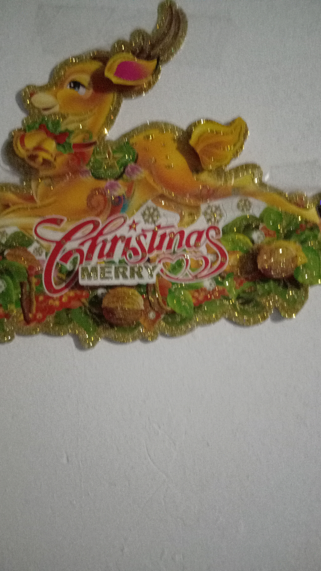 The image shows a Christmas decoration hanging on a light-colored wall. The decoration is shaped like a reindeer with the words "Merry Christmas" written on it. The reindeer is yellow and has glittery antlers and ears. The words are in red and white with gold accents. The decoration is surrounded by green foliage and brown ornaments. The wall is plain and there is nothing else visible in the image. The location is unspecified.
