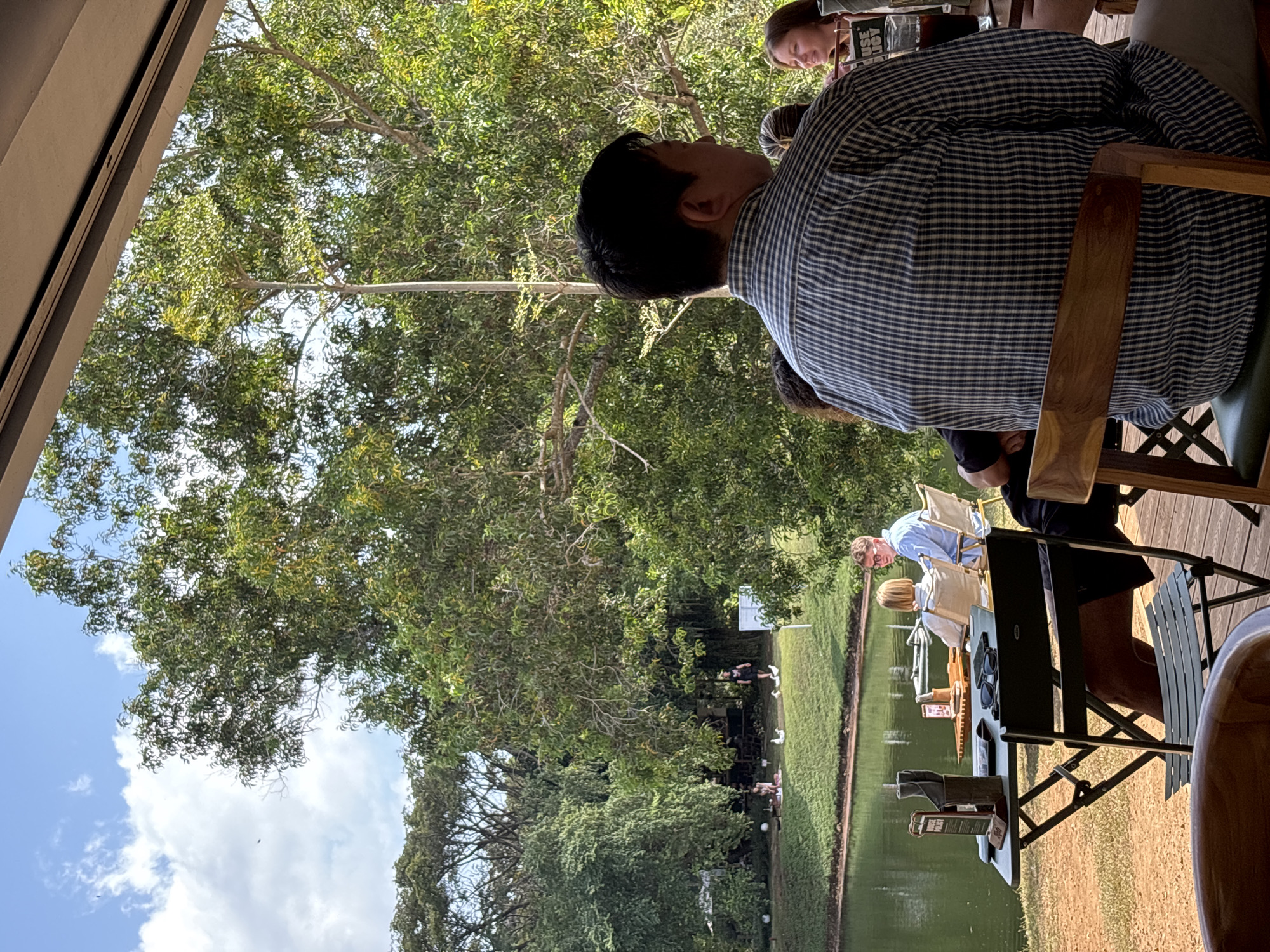 The image captures an outdoor dining or relaxation setting, offering a scenic view of a lush landscape. The perspective appears to be from a raised wooden deck or patio, looking outwards over a natural environment.
In the mid-ground, a man with dark hair is prominently seated with his back to the viewer, wearing a blue and white checkered shirt. He occupies a wooden chair with a dark green cushion. Behind him, another person is partially visible, holding what appears to be a book or menu that displays the text "USE JOY". On a table near them, a pair of sunglasses is placed.
The scene is dominated by abundant green foliage from numerous large trees, suggesting a tropical setting. Beyond the immediate seating area, a calm body of water, likely a pond or small lake, stretches across the frame, with a manicured grassy bank on the opposite side. Further in the distance, more people are gathered, some seated at tables with white umbrellas, and others standing near the water's edge, where small white objects are visible on the grass. One individual in the background is wearing a light blue shirt.
The upper left corner of the image reveals a bright blue sky with scattered white clouds, indicating a clear, sunny daytime. The overall ambiance is tranquil and inviting, suitable for a leisurely meal or relaxation in nature. This serene outdoor environment is located in Kuta Selatan, Indonesia.