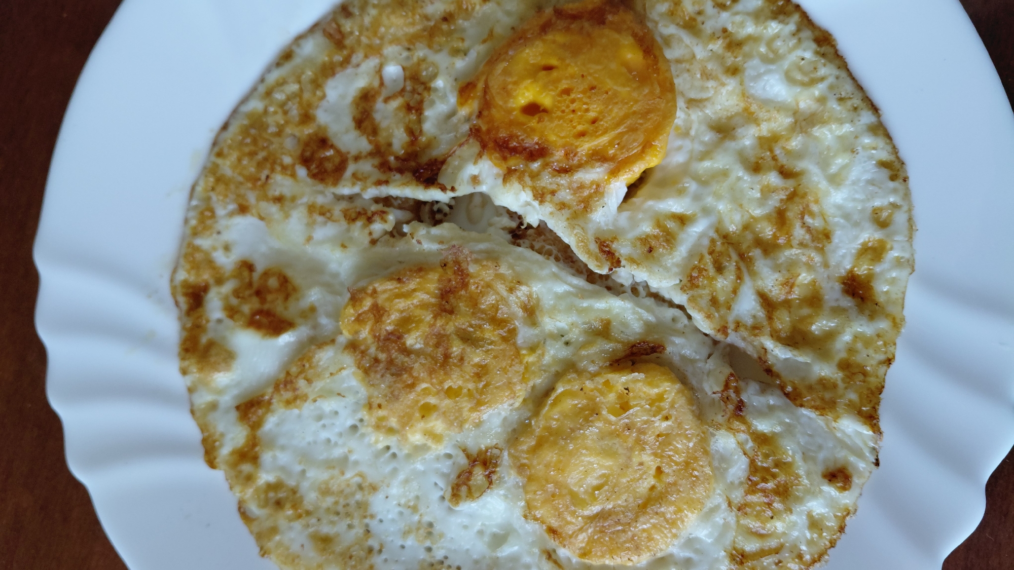 A close-up photograph shows two fried eggs on a white plate. The egg whites are cooked, slightly browned around the edges, and the yolks are also cooked, displaying a firm, orange-yellow color. The plate is plain white and appears to be sitting on a brown surface, possibly a wooden table. The setting is Unknown, Unknown.