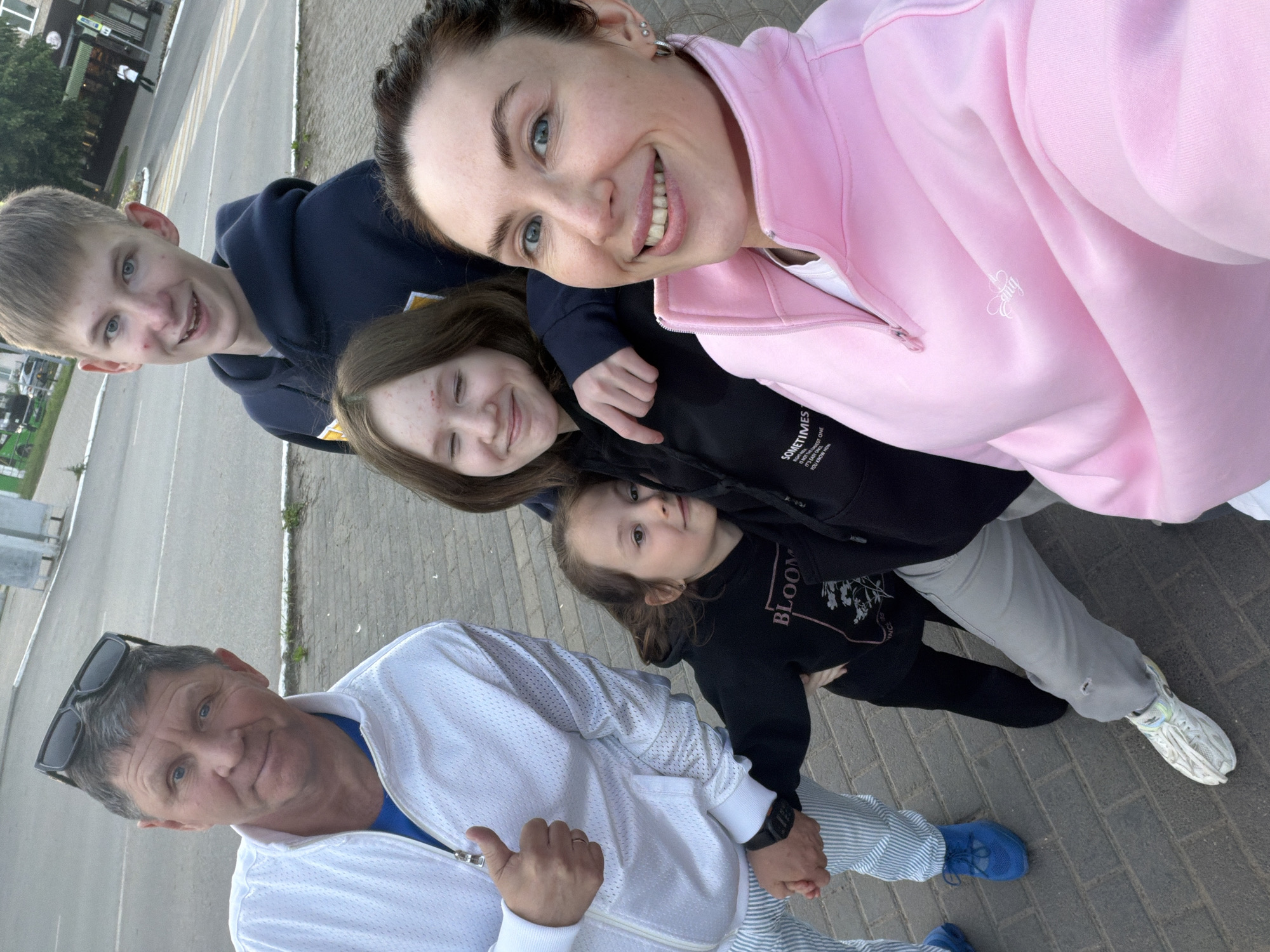 The image captures a group selfie of five people, presumably a family, outdoors in Chaikovsky, Russia. An adult woman with a wide smile, wearing a pink zip-up jacket, holds the camera. Faint white embroidery, possibly "Hug," is visible on her jacket. To her left, a smiling teenage boy in a dark blue hoodie looks towards the camera. Below the woman, a teenage girl in a black hoodie with small, partially visible white text ("SOMETIMES IT'S THE...") grins. Next to her, a young girl in a black hoodie featuring "BLOOM" and floral graphics looks calmly at the lens. At the bottom, an adult man with sunglasses resting on his head wears a white mesh-like zip-up jacket and vertically striped pants, smiling. He holds hands with the young girl.
They are standing on a paved surface, a combination of brick pavers and asphalt, likely a sidewalk adjacent to a road with a visible crosswalk in the background. Buildings and some trees line the distant street. The scene is illuminated by soft daylight, suggesting a pleasant daytime setting. All individuals exhibit happy and relaxed expressions, conveying a sense of family togetherness. The man wears blue sneakers, and the young girl has white sneakers.