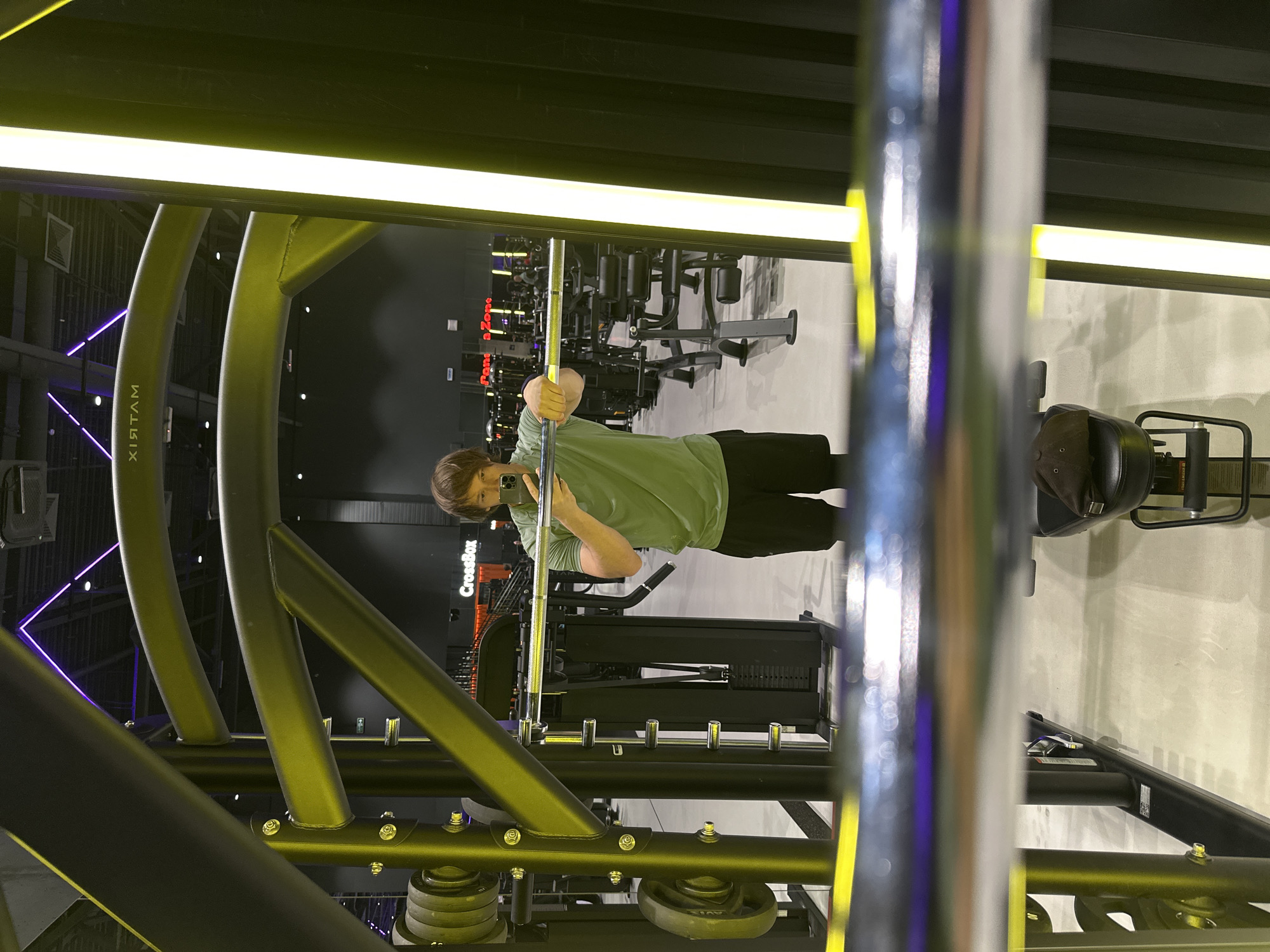 A man wearing a green t-shirt and black shorts is taking a selfie in a gym. He is holding a barbell with both hands. The gym appears to be a modern facility with specialized workout equipment, including weight machines and racks. The lighting is predominantly yellow and purple, with prominent vertical light strips. In the background, a sign reads "CrossBox". The floor is light-colored and reflective, showing reflections of the gym equipment and lights. The man is positioned centrally in the frame, with the gym equipment forming a metal structure around him, partially obscuring him and the environment. The overall scene suggests a workout session in progress. The image also shows a cap on a piece of gym equipment in the foreground. The location is identified as Murino, Russia.