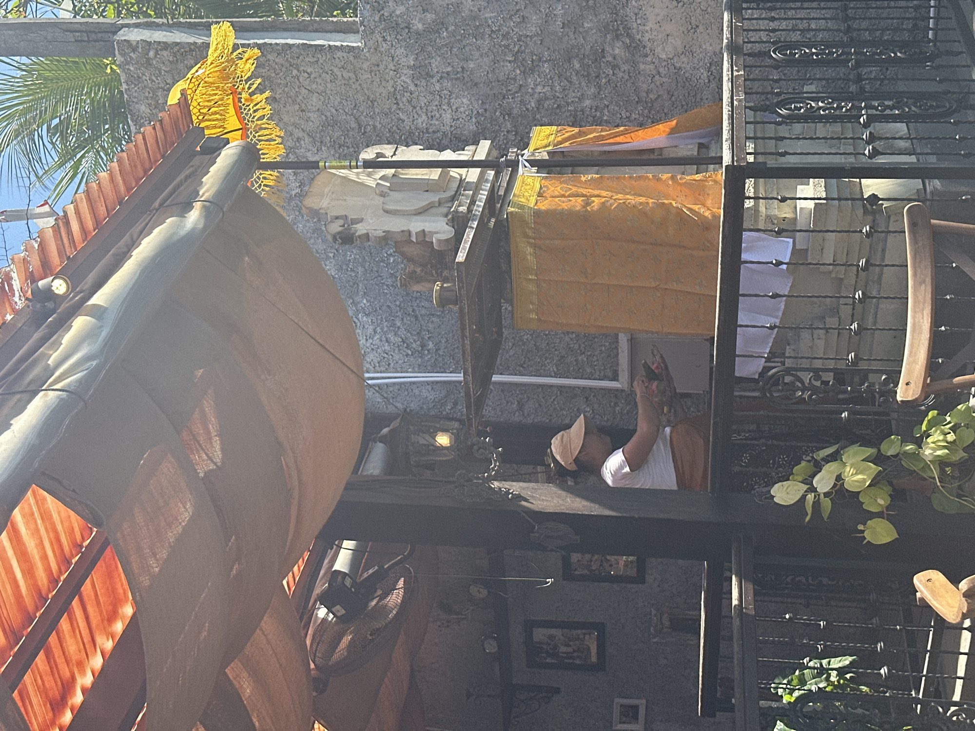 A Balinese woman wearing a white t-shirt and a straw hat is standing on a balcony in Denpasar, Indonesia. She is holding a tray of offerings, likely for a religious ceremony. Behind her, a stone statue is draped with an orange cloth. The balcony is covered by a straw roof and decorated with a yellow fringe. The scene appears to be a temple or a place of worship.