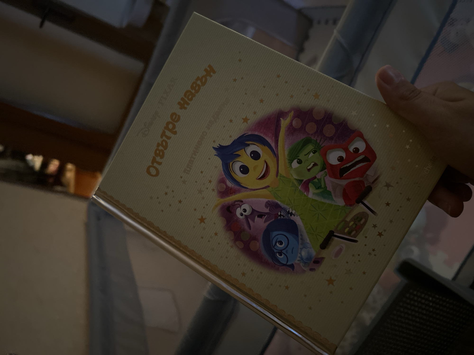 The image shows a close-up of a book held by a person's hand. The book has a light yellow cover with an illustration of characters from the Disney Pixar movie "Inside Out." The characters depicted are Joy, Sadness, Fear, Disgust, and Anger. The title of the book appears to be in Bulgarian script, reading "Отвътре навън" (which translates to "Inside Out"). The background of the image is out of focus and appears to be an indoor setting, possibly a room with furniture, but no specific city or location details are discernible from the image itself.