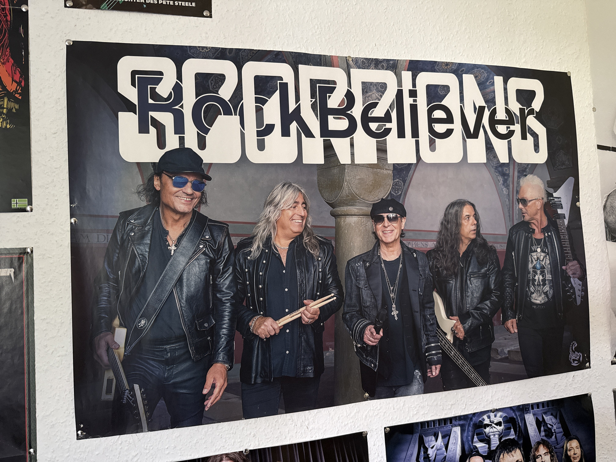 In Bad Sachsa, Germany, a poster of the rock band Scorpions is displayed on a textured white wall. The poster features the band's logo, "ROCK BELIEVERS," in large, white, block letters with a dark outline. Below the logo, five band members are pictured. From left to right, the first member wears a black baseball cap, sunglasses, and a black leather jacket. The second member has long, grey hair, a black collared shirt adorned with studs, and is holding drumsticks. The third member, in the center, has long, dark hair and a black leather jacket. The fourth member, on the right, has spiky blonde hair, sunglasses, a black leather jacket, and is holding a white electric guitar. The fifth member, partially visible on the far right, is also wearing a black leather jacket and holding a microphone. The background of the poster depicts an architectural setting with arches and patterned wallpaper. Below the Scorpions poster, another poster is partially visible, featuring imagery related to the band Iron Maiden. The wall is also adorned with smaller posters and what appear to be thumbtacks or pins holding them in place.