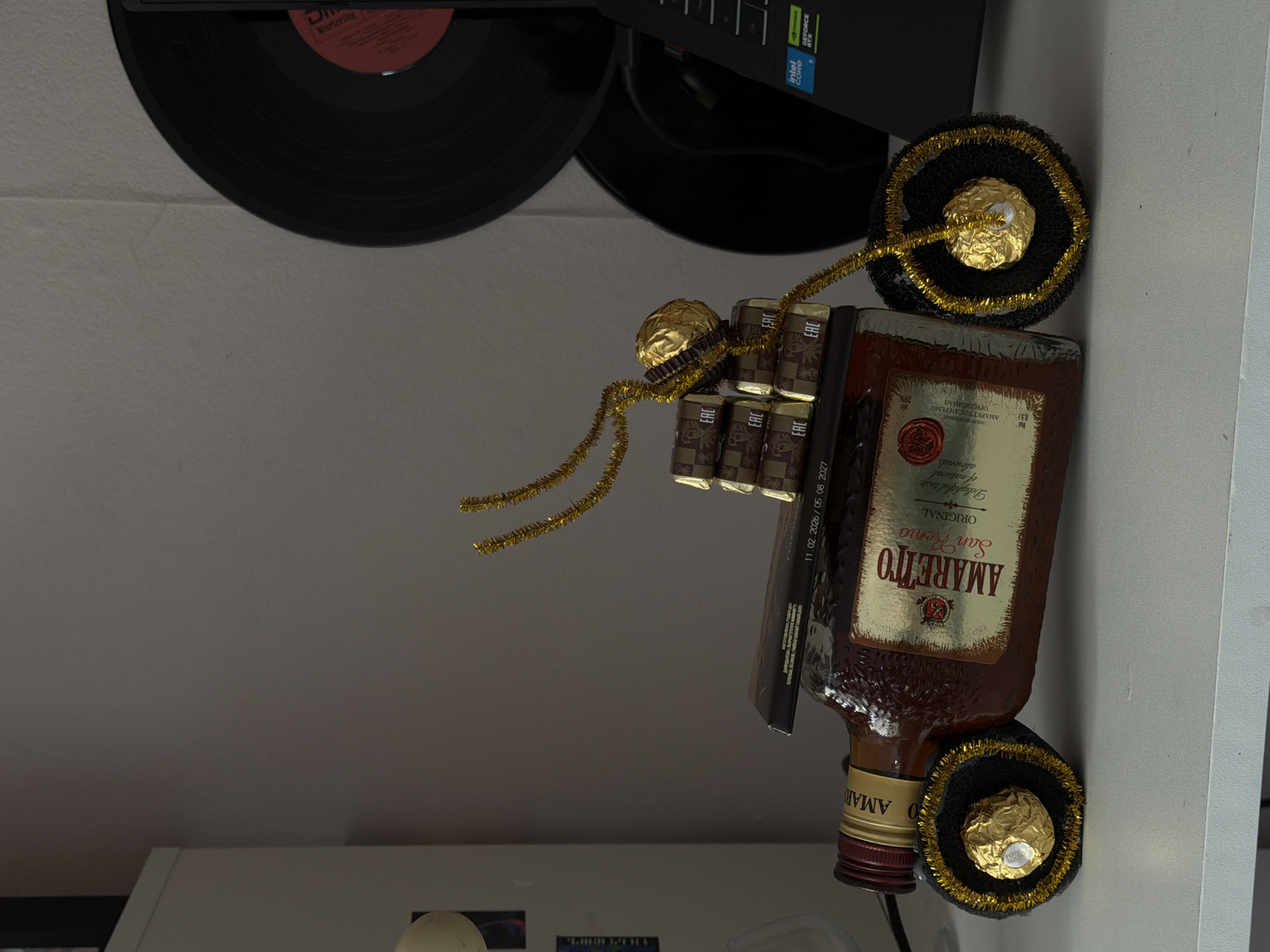 An indoor still life arrangement, rotated 90 degrees clockwise, displays a bottle of Amaretto liqueur and Ferrero Rocher chocolates, possibly on a desk or shelf. The prominent subject is a horizontal bottle of "AMARETTO San Remo ORIGINAL," with its label detailing "Delightful taste of natural almonds," "Vol 0,51," and "28%" alcohol content. Cyrillic text on the label reads "ЛИКЕР "АМАРЕТТО САН РЕМО" ОРИДЖИНАЛ" and mentions "ИЗГОТОВЛЕНО И РАЗЛИТО ПО СПЕЦИАЛЬНОМУ ЗАКАЗУ."
Decorative elements include several gold-foiled Ferrero Rocher chocolates and black and gold tinsel. Two chocolates are encased in black, tinsel-edged circular bases at either end of the bottle, while a cluster of four chocolates rests atop the bottle, adorned with dangling gold tinsel. A black box positioned next to the bottle shows production/expiration dates "11.02.2026 / 05.08.2027." "EAC" marks are visible on the chocolate wrappers and the bottle.
In the background, two partially visible black vinyl records are present, one showing "Mastering" on its label. Above these, a black electronic device, likely a computer component, features "intel CORE" and "GEFORCE RTX" logos. The scene is well-lit, characteristic of an indoor environment. The arrangement suggests a decorative display or a gift.