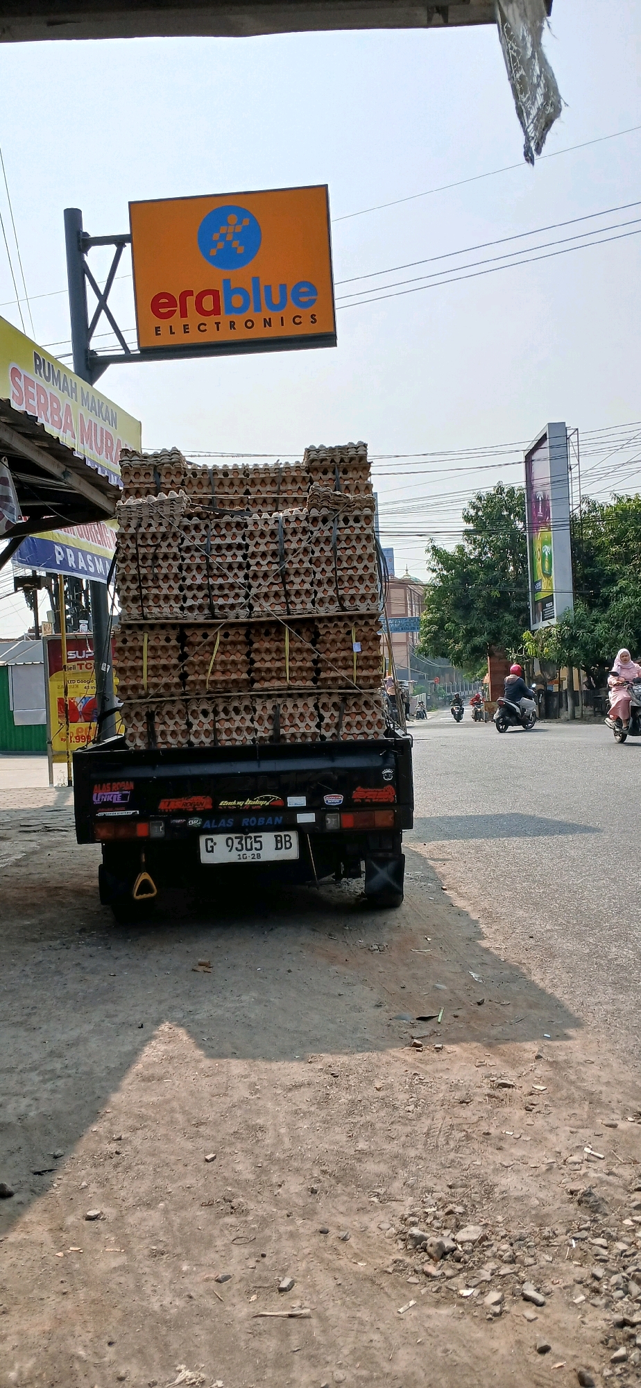 The image shows a black pickup truck parked on the side of a road in Cirebon, Indonesia, loaded with a large stack of egg cartons. The truck has a license plate that reads "G 9305 BB". In the background, there is a sign for "erablue ELECTRONICS" and a yellow sign with Indonesian text that includes "RUMAH MAKAN SERBA MURAH". There are trees and buildings visible along the road, and several motorcycles and a scooter with riders are on the road. The sky is overcast. The ground in the foreground is dusty and uneven.