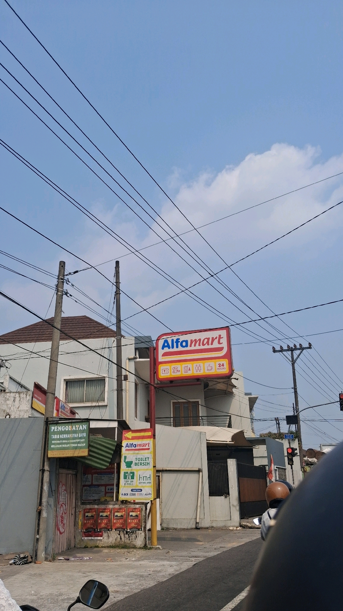 The image depicts a street scene in Tlatar, Indonesia, under a clear blue sky with scattered clouds. Overhead, a dense network of power lines crisscrosses the frame, creating a visually complex canopy. In the mid-ground, a two-story building with a brown tiled roof is visible, partially obscured by the power lines. Dominating the right side of the building is a prominent red and white Alfamart sign, indicating a retail store. Below the Alfamart sign, a yellow directional sign with the Alfamart logo points to a "TOILET BERSIH" (clean toilet), "TOP-UP E-TOLL," and a sitting area.
To the left of the Alfamart, a separate entrance is visible with a sign above it that reads "PENGOBATAN TABIB HERMAN SYAH BAYU" (Medical Treatment by Tabib Herman Syah Bayu). This entrance is adorned with posters. In front of this entrance, a small green corrugated metal roof is propped up.
Further to the right, a traffic light shows a red signal. A motorcyclist wearing a brown helmet and a light-colored jacket is partially visible in the foreground, suggesting movement or a pause in traffic. The street in the foreground is asphalt. The overall impression is a snapshot of a commercial area in an Indonesian city.