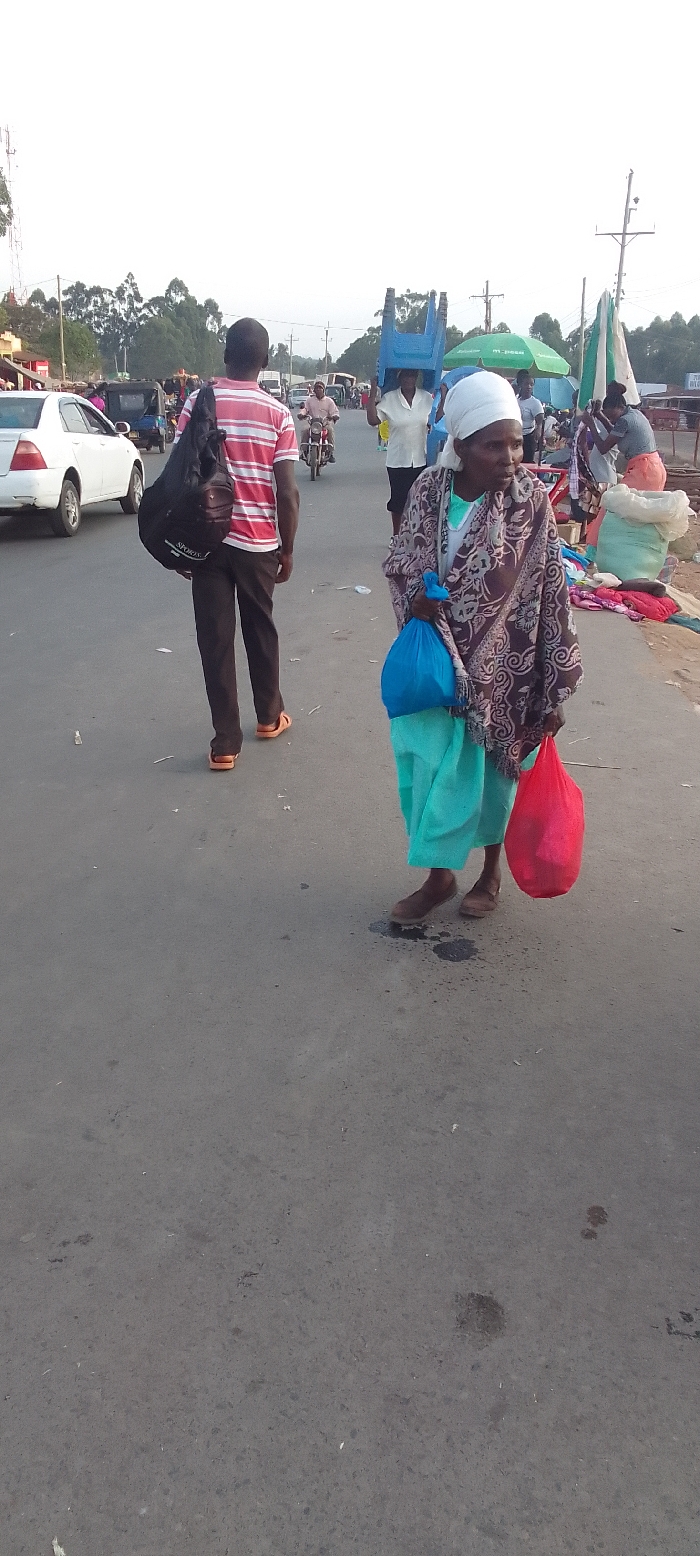 The image depicts a bustling street scene in Kakunga, Kenya, likely in the late afternoon or early morning due to the soft light. In the foreground, an elderly woman walks towards the right, wearing a white headscarf, a patterned brown and white shawl over a light blue skirt, and simple dark shoes. She carries a bright blue plastic bag in her left hand and a red plastic bag in her right. Her expression appears neutral or contemplative.
Behind her, a man with his back to the camera walks away, wearing a red and white striped t-shirt, dark trousers, and orange sandals. He carries a black shoulder bag that has the word "SPORTS" visible on it.
The street is an asphalt road with scattered debris. In the background, there's ongoing activity typical of a market or busy town center. A person is seen carrying a blue plastic chair on their head. Other pedestrians, motorcycles, and a white car are visible, indicating shared road usage. Market stalls with green and white umbrellas are present on the right side of the road, and sacks of goods are visible near them. One green umbrella clearly displays "AIRTEL MONEY" and phone numbers starting with "070..." and "078...". The scene is framed by trees and utility poles in the distance, under a bright, overcast or hazy sky.