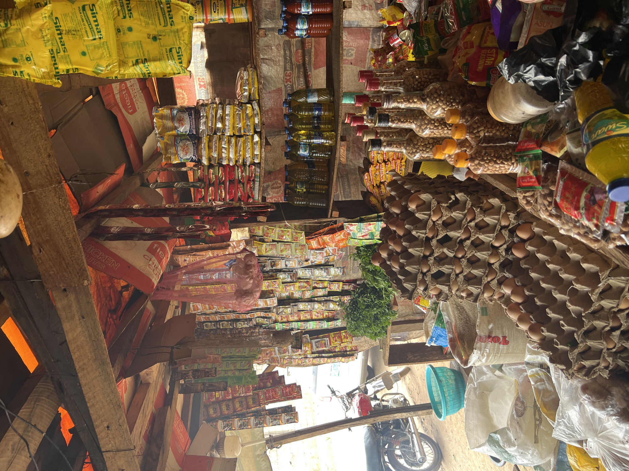 This image captures a vibrant and densely packed market stall or small shop, likely in Abuja, Nigeria. The scene is an indoor commercial space, characterized by an abundance of various food products displayed on makeshift shelves fashioned from wooden planks and cardboard boxes. No people are visible, but the arrangement of goods clearly indicates a retail setting where items are prepared for sale.
The stall is overflowing with merchandise. On the upper left, large yellow bags of "Simas" margarine (with text in English, French "POUR CUIRE ET CUISINER", and Portuguese "ARISAR E COZINHAR") are prominently displayed. Below them, numerous small sachets of seasonings and instant noodles (some marked "fiesta CHICKEN GUTS") are stacked. Various bottles containing cooking oil, including "Swan" brand, and plastic bottles filled with peanuts, are arranged neatly on wooden shelves in the center. Rows of eggs are carefully stacked in cardboard cartons on the right side of the image, while bags of grains or flour, one of which reads "Total," rest on the floor. Other visible items include packets of "MAGGI SEASONING," "STAR SEASONING," "Curry Powder," and products from the "Gino" brand. A small pile of fresh green leafy vegetables is also present.
Natural light appears to enter from outside, illuminating parts of the interior. Outside the stall, a motorcycle is partially visible, indicating a bustling street or market environment. The overall impression is one of a busy, local provisions shop, typical of street markets in West Africa, efficiently utilizing every available space to display its wide array of goods.
