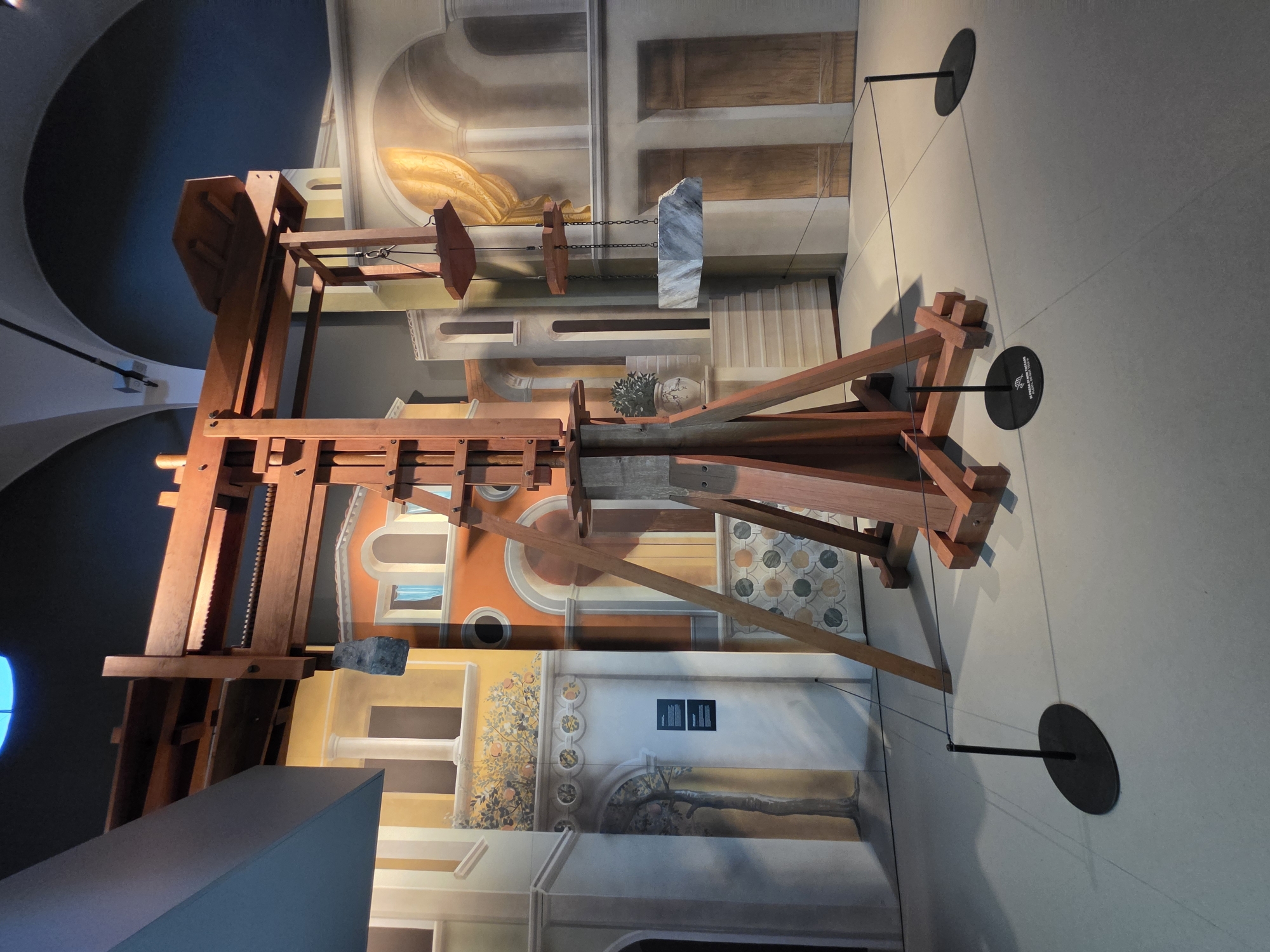 The image shows a wooden replica of a Leonardo da Vinci crane, displayed in an exhibition space. The crane is positioned in the center of the frame, with a large piece of marble suspended from it by chains. The background is a mural depicting an Italian Renaissance cityscape with buildings, arches, and a draped curtain. There are no people visible in the image. The exhibition space appears to be indoors, with a high, arched ceiling visible in the upper part of the image. Black stanchions with ropes are placed around the exhibit to keep visitors at a distance. The flooring is a light grey, smooth surface. The location is identified as the Museo della Scienza e della Tecnologia "Leonardo da Vinci" in Milan, Italy, which is a science museum dedicated to Leonardo da Vinci's work.