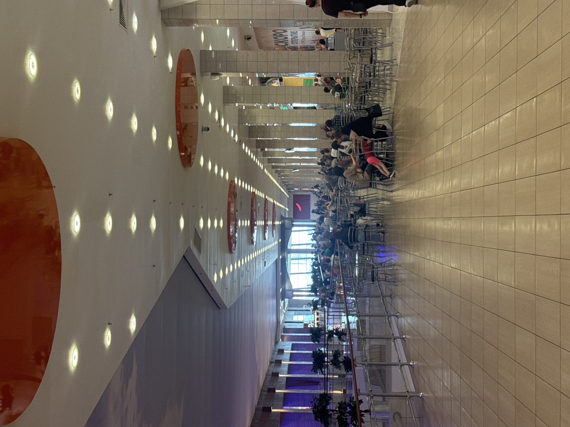 This image depicts a wide view of a modern indoor space, likely a food court or common area within a shopping mall or similar establishment in Saint Petersburg, Russia. The scene is characterized by its long, linear layout, with one side featuring a white, slightly curved wall adorned with numerous circular, illuminated lights. On this wall, two large, orange, oval-shaped decorative elements are prominently displayed.
The opposite side of the space is lined with rows of metal chairs and tables, suggesting a seating area. A significant number of people are seated at these tables, engaged in various activities, possibly dining or socializing. The crowd appears diverse in age and attire. The flooring consists of large, rectangular beige tiles, reflecting the overhead lighting.
Further down the corridor, a large window or open area is visible, admitting natural light and offering a glimpse of what appears to be an outdoor or more open space beyond. This area also has a tiered, stepped structure with blue lighting, adding a modern aesthetic. A banner with text is visible in the background above the seating area. The lighting in the scene suggests it could be daytime, as indicated by the mix of artificial and natural light. The overall impression is one of a busy, contemporary public space.