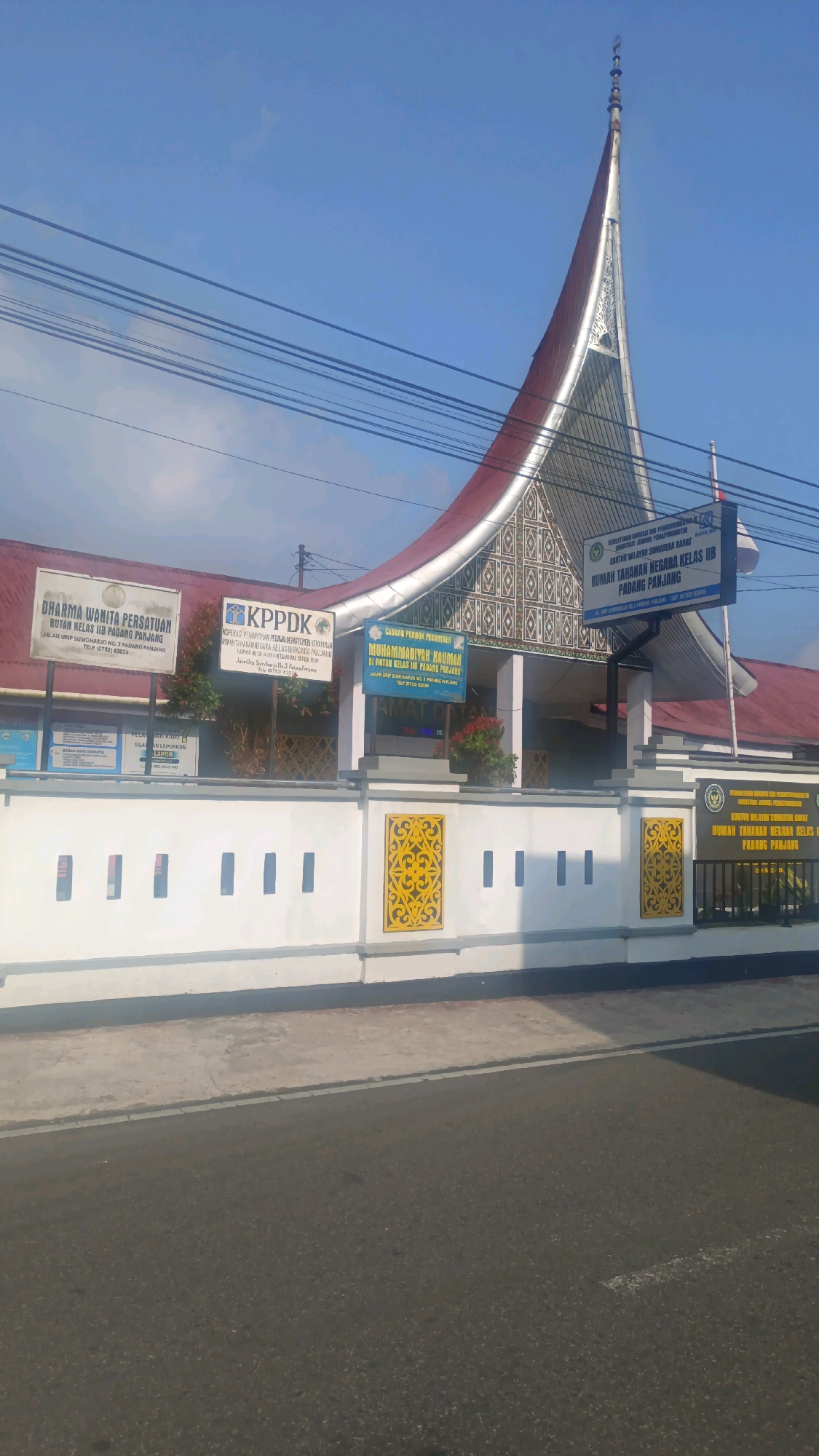 The image depicts the front facade of a building in Padang Panjang, Indonesia, identified by signage as RUMAH TAHANAN NEGARA KELAS II B PADANG PANJANG. The building features a distinct, ornate roof with a pointed, curved design, reminiscent of traditional Minangkabau architecture. In front of the building is a white wall adorned with decorative yellow patterns. Several signs are visible, including one that reads "DHARMA WANITA PERSATUAN ROTAN KELAS II B PADANG PANJANG" and another with "KPPDk". The building appears to be operational, with electrical wires visible overhead and a clear blue sky in the background. The scene is set on a street, suggesting a public area. No people are visible in the image.