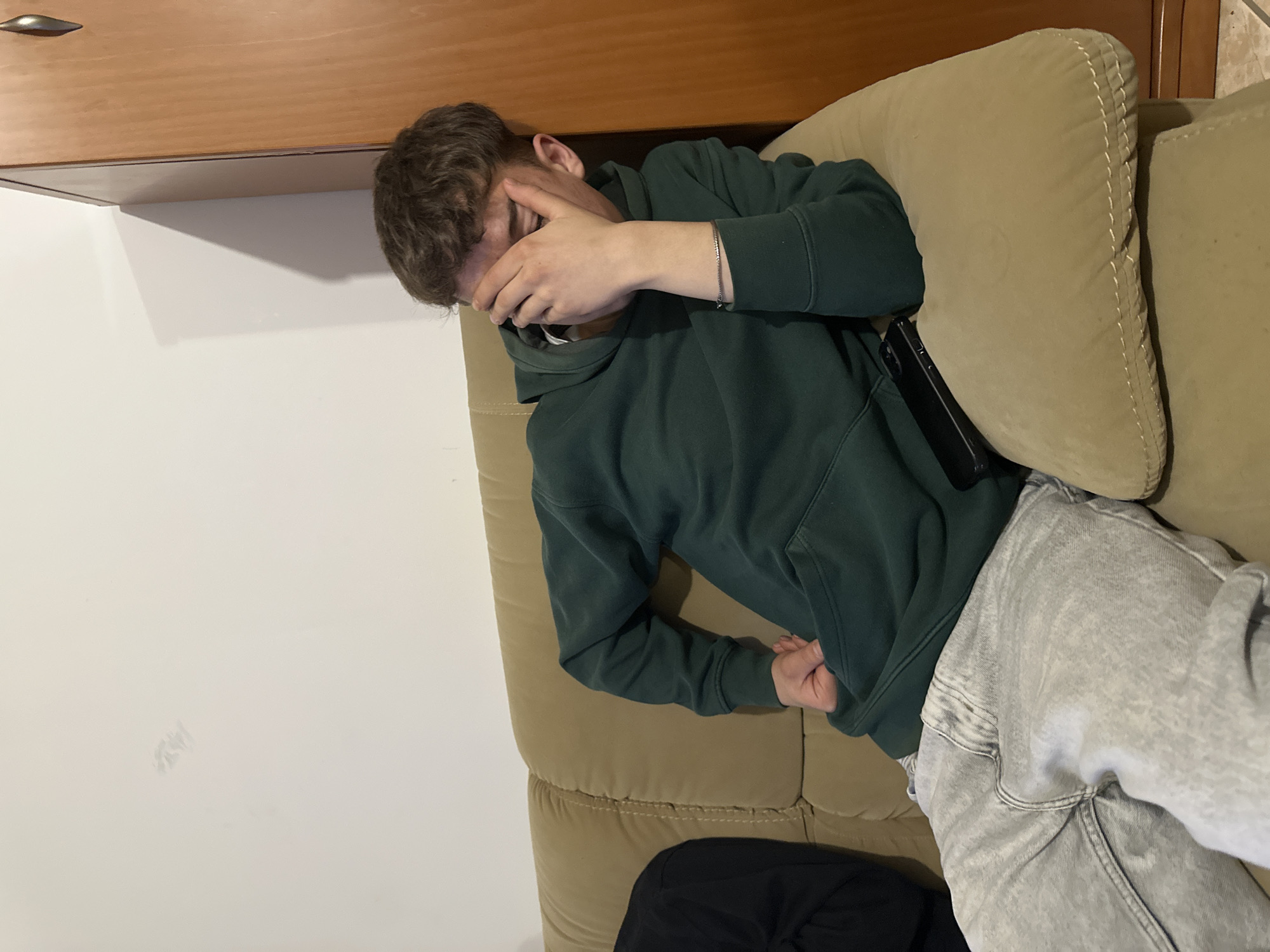 A young man is lying on a tan-colored sofa in an indoor environment. He is wearing a dark green hoodie and light gray jeans. His right hand is covering his face. A black mobile phone is visible on the sofa next to him. The sofa is positioned against a white wall and a wooden cabinet. There is no indication that this image was taken in Bragança, Portugal.