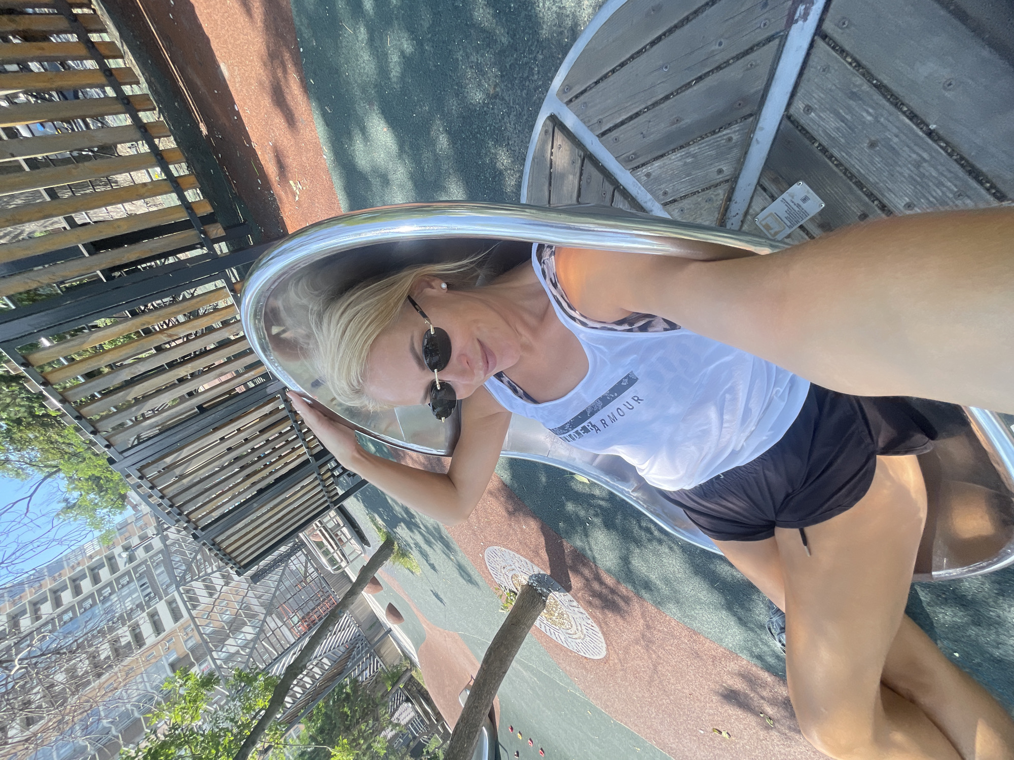 A blonde woman is sitting on a curved metal slide in a park. She is wearing a white tank top with leopard print straps, black shorts, and sunglasses. She is looking at the camera and has her left hand on the slide. The park has a rubberized surface with red and green sections, trees, and a wooden fence in the background. In the distance, a tall building can be seen. The image appears to be taken in an urban park on a sunny day.