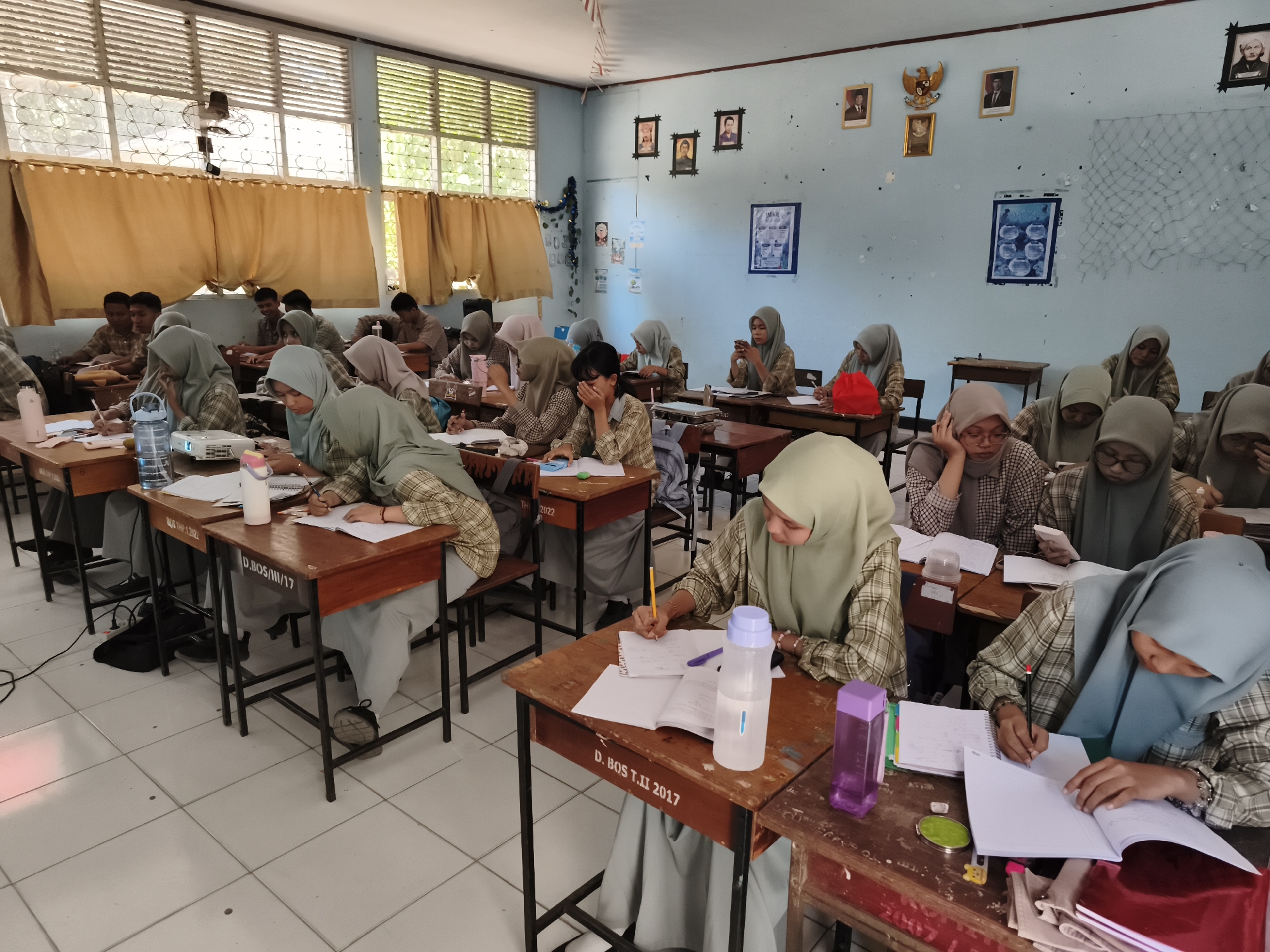 This image depicts a classroom in Makassar, Indonesia, likely a school setting, with a group of students, predominantly female, seated at desks. The students are dressed in uniforms, many wearing hijabs. They appear to be engaged in learning activities, with some writing in notebooks and others looking at their materials. The classroom environment shows typical school furniture, including wooden desks and chairs, and the walls are adorned with framed pictures and educational posters. Sunlight streams in through the windows, illuminating the scene. The overall atmosphere suggests a regular school day in progress.