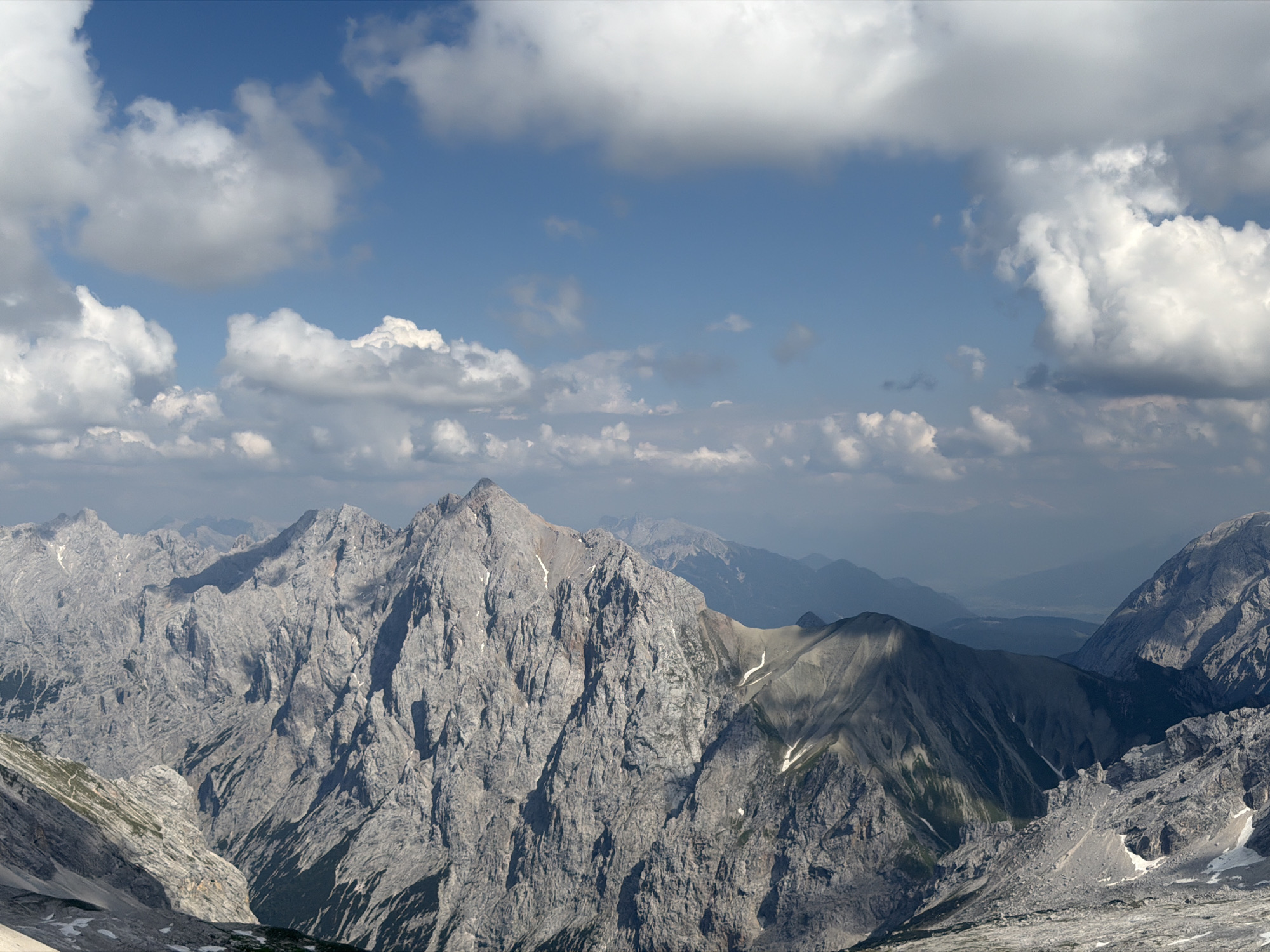 This image depicts a panoramic view of rugged, gray mountain peaks under a bright blue sky dotted with fluffy white clouds. The mountains are steep and rocky, with some patches of snow visible on their slopes. The lighting suggests it is daytime, likely midday or early afternoon, with the sun casting shadows that highlight the contours of the landscape. The scene conveys a sense of vastness and natural beauty. In the distance, other mountain ranges fade into a hazy blue, indicating a considerable distance and depth to the landscape. The overall atmosphere is one of serene, alpine wilderness. No people, objects, or text are visible in the image.