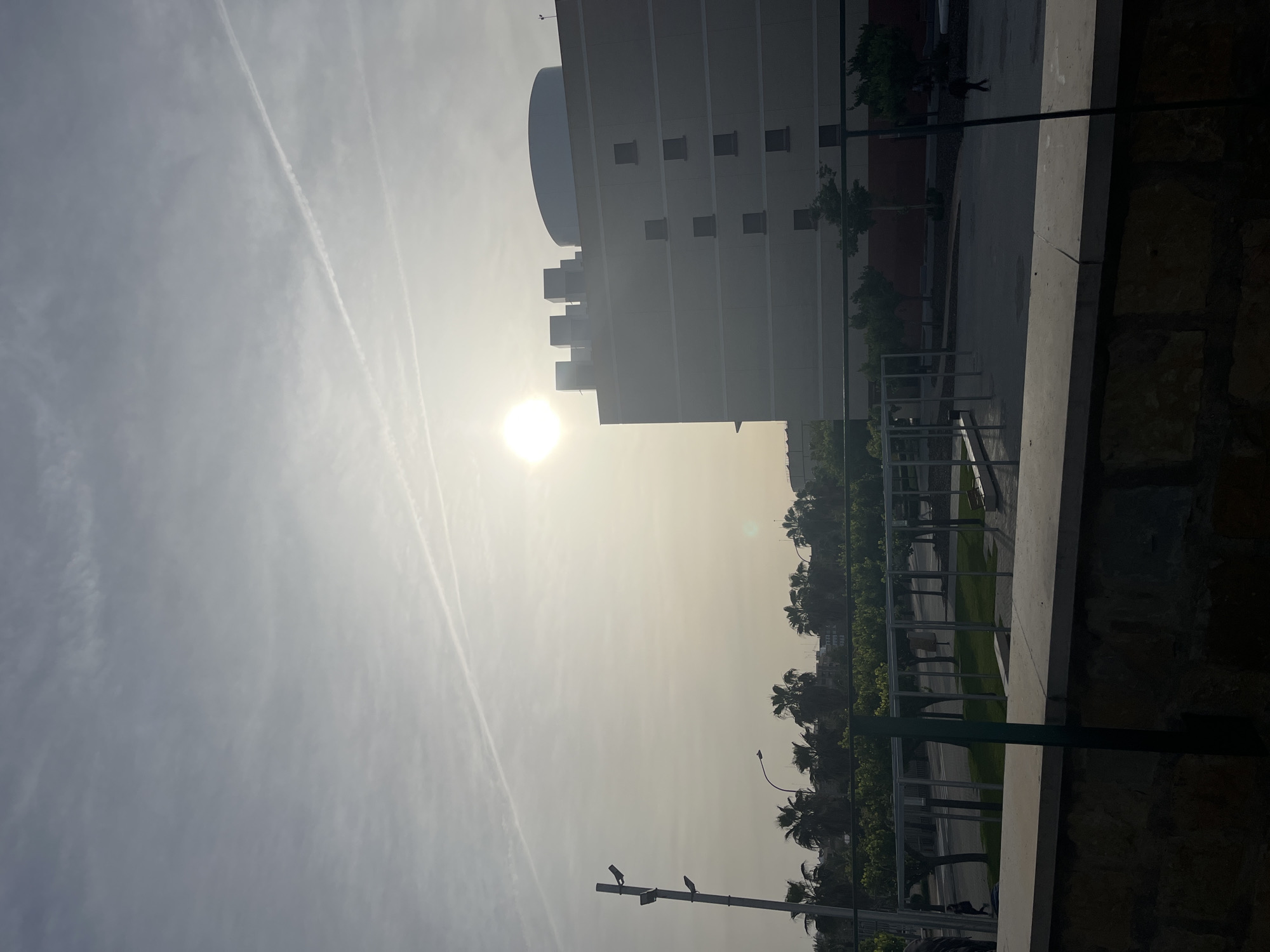 The image, presented with a 90-degree counter-clockwise rotation, depicts a bright, hazy outdoor scene under intense sunlight. When viewed in standard orientation (rotated 90 degrees clockwise), a brilliant, almost overexposed sun dominates the upper-left quadrant of the light grey, hazy sky. Several distinct contrails from aircraft are visible streaking across the sky in the upper-left and central areas.
A large, multi-story building with light-colored facades and numerous small, rectangular windows occupies the middle-right and upper-right portions of the frame. Its rooftop features a prominent, large rounded structure, possibly a dome or tank, alongside several smaller, blocky architectural elements.
The lower half of the scene shows a paved ground area interspersed with dark green foliage and trees, including several tall palm trees, suggesting a warm climate consistent with València, Spain. A dark, slender streetlight pole stands in the lower-left corner. In the background, near the building and trees, two small, dark, indistinct figures of people can be seen walking or standing.
In the immediate foreground, on the right side, there is a rough stone wall or barrier, topped by a smooth concrete ledge. A dark metal railing system, including a thick dark green vertical support post, is also visible in the lower-right foreground.
The intense, direct sunlight indicates a daytime scene, likely midday or early afternoon, under bright yet somewhat hazy weather conditions. No discernible text is visible in the image.