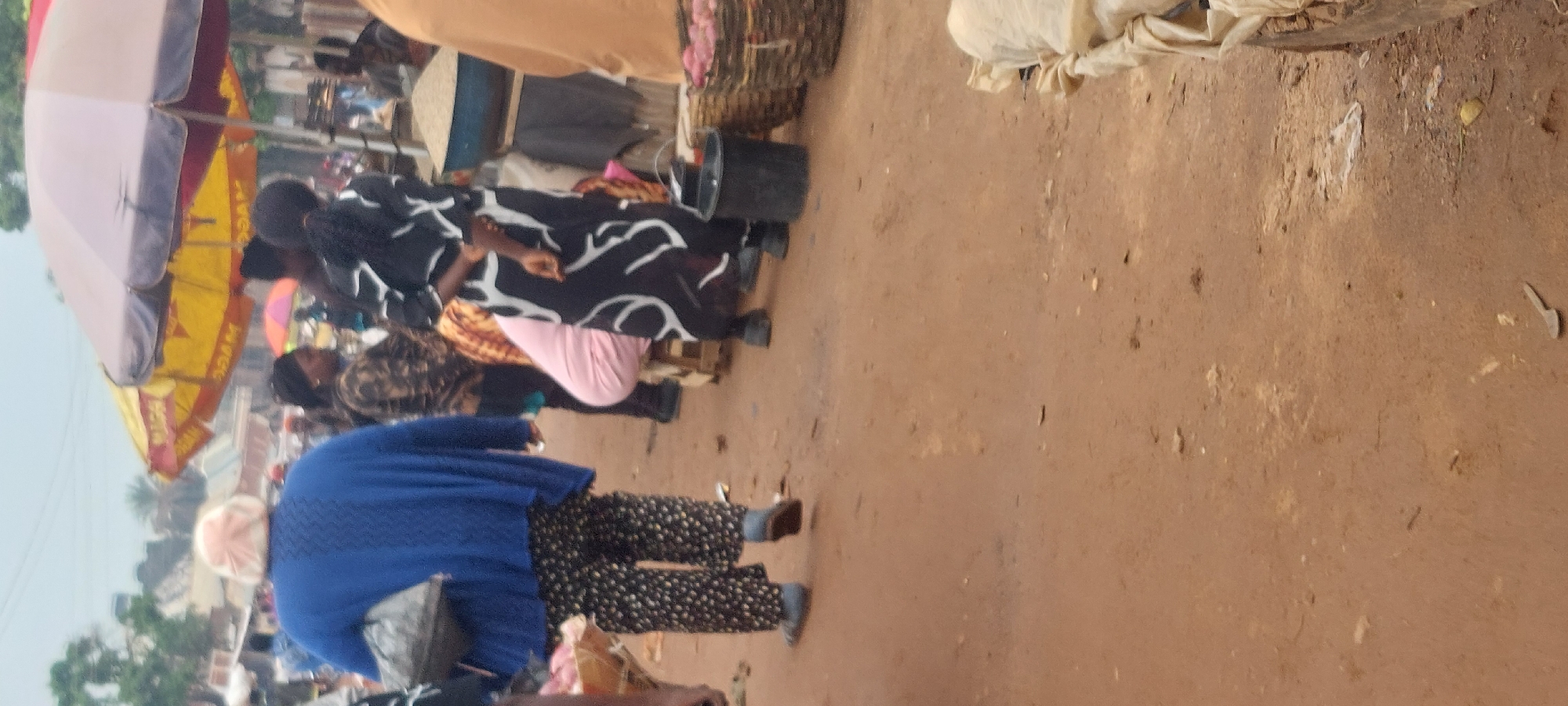 In an outdoor market, a person in a blue sweater and patterned pants walks away from the camera, carrying a black bag. Another person in a black and white patterned dress stands with their back to the camera, with a person in a pink garment crouched in front of them. Several stalls are visible, with umbrellas providing shade, and baskets containing produce. The ground is dirt and scattered with debris.