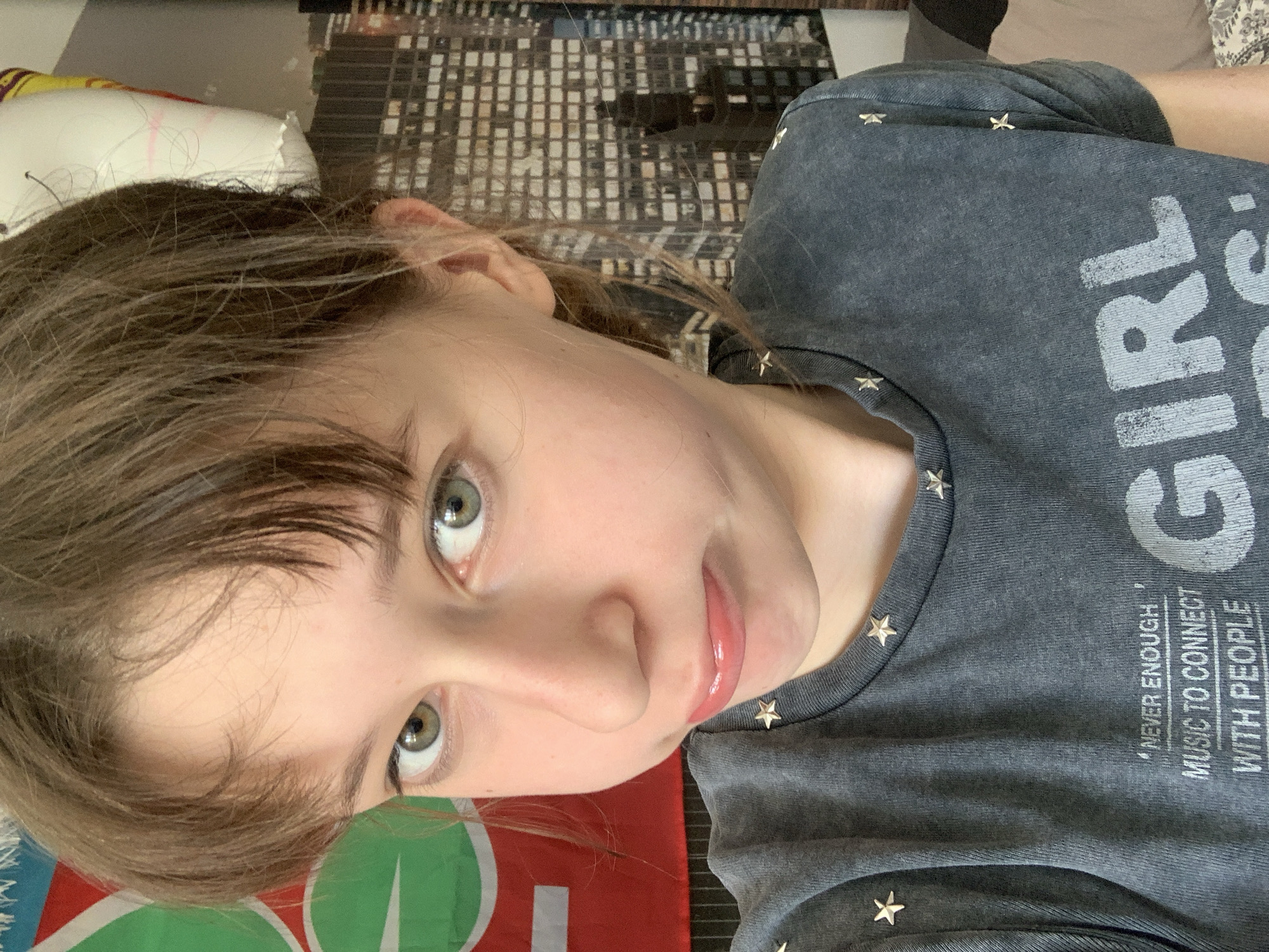 A young person with fair skin and light brown hair is facing the camera. They are wearing a dark gray t-shirt with a graphic that includes the words "GIRL" and "NEVER ENOUGH MUSIC TO CONNECT WITH PEOPLE," and adorned with small star-shaped studs. The person is lying on a surface with a patterned background showing a cityscape. The setting is indeterminate, but is not located in Vozdovac Urban Municipality, Serbia.