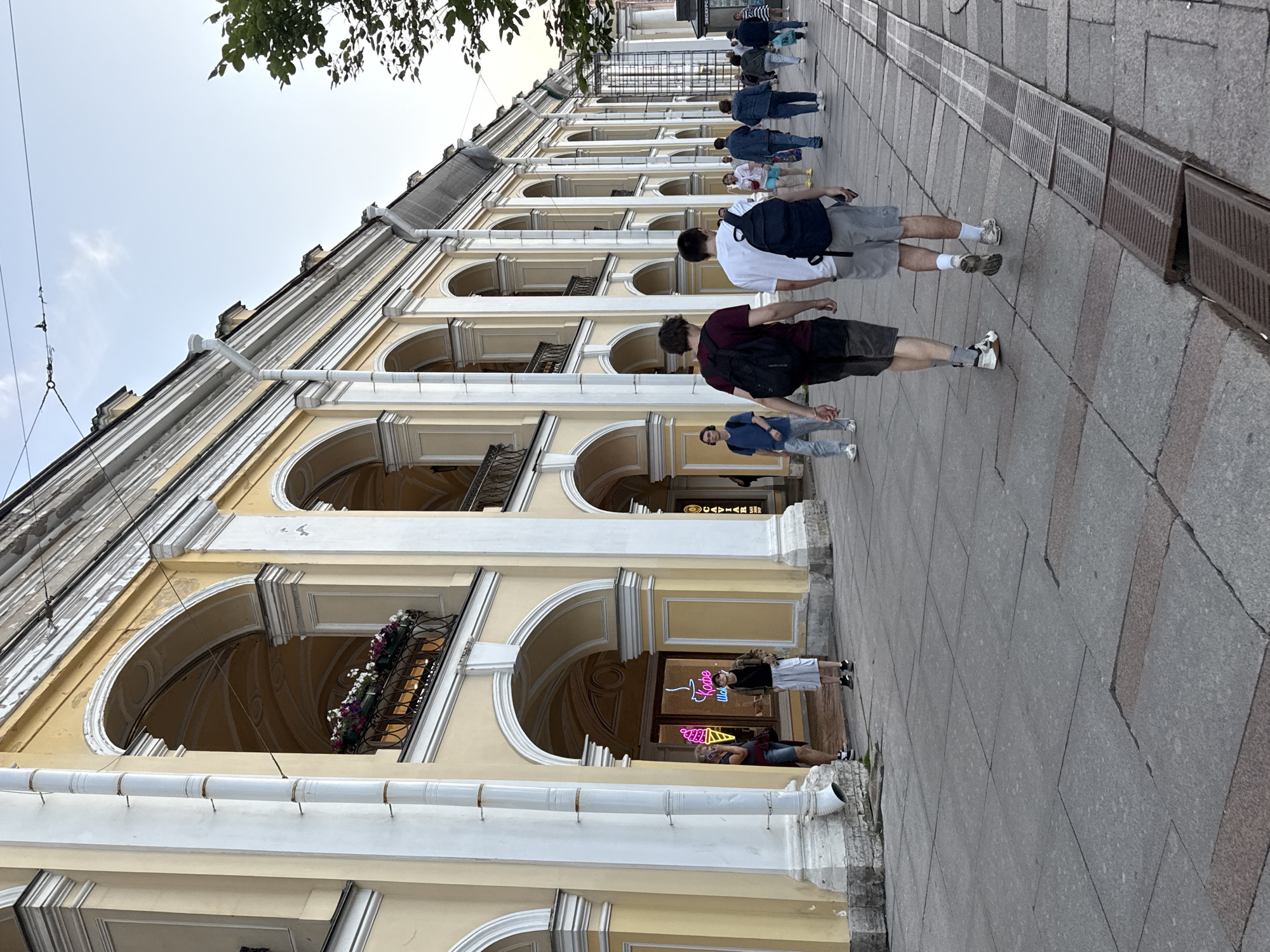 The image captures a view of the Gostiny Dvor arcade in Saint Petersburg, Russia, with pedestrians walking along the sidewalk in front of the yellow-colored building. The arcade features a series of arched openings and balconies adorned with flower boxes. Two young men with backpacks are walking away from the camera, and other individuals are scattered throughout the scene. A neon sign for an ice cream shop is visible through one of the archways. The ground is paved with large stone slabs, and a metal grate runs along the edge of the walkway. The sky is overcast, suggesting either early morning or late afternoon.