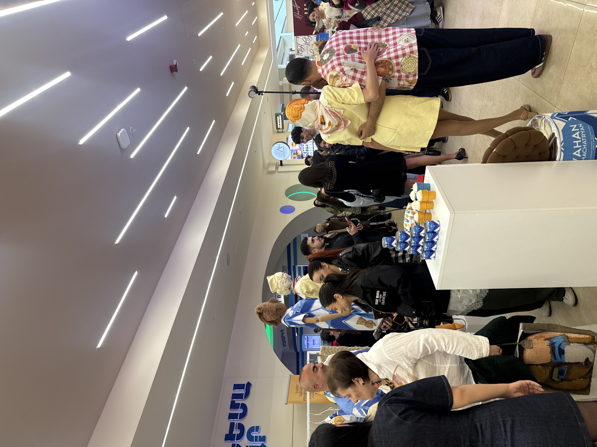 A queue of people stands in what appears to be a shopping mall in Yerevan, Armenia. The scene is illuminated by long, linear ceiling lights. A man in a red and white plaid shirt is standing next to a woman in a yellow dress and an orange headscarf. Several other individuals are lined up behind them, some holding phones. A display of beverages is visible on a white table. Armenian text is visible on signage in the background.
