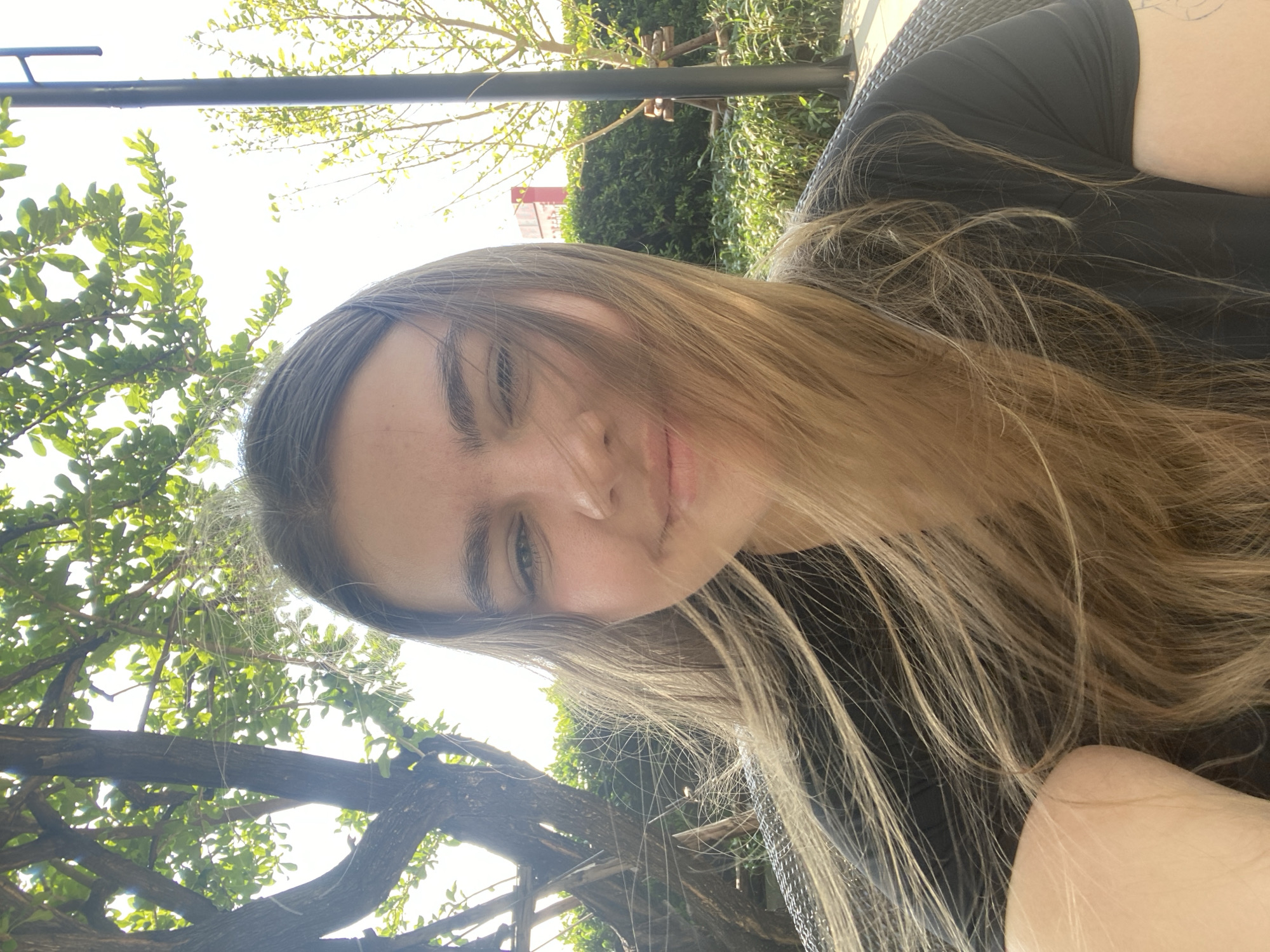 A young woman with long, highlighted blonde hair is taking a selfie outdoors. She is wearing a dark-colored top. The background shows a green leafy tree canopy and a metal support structure overhead. The presence of tropical foliage suggests a warm climate. Without further information, it's impossible to verify if the location is Bangkok, Thailand.