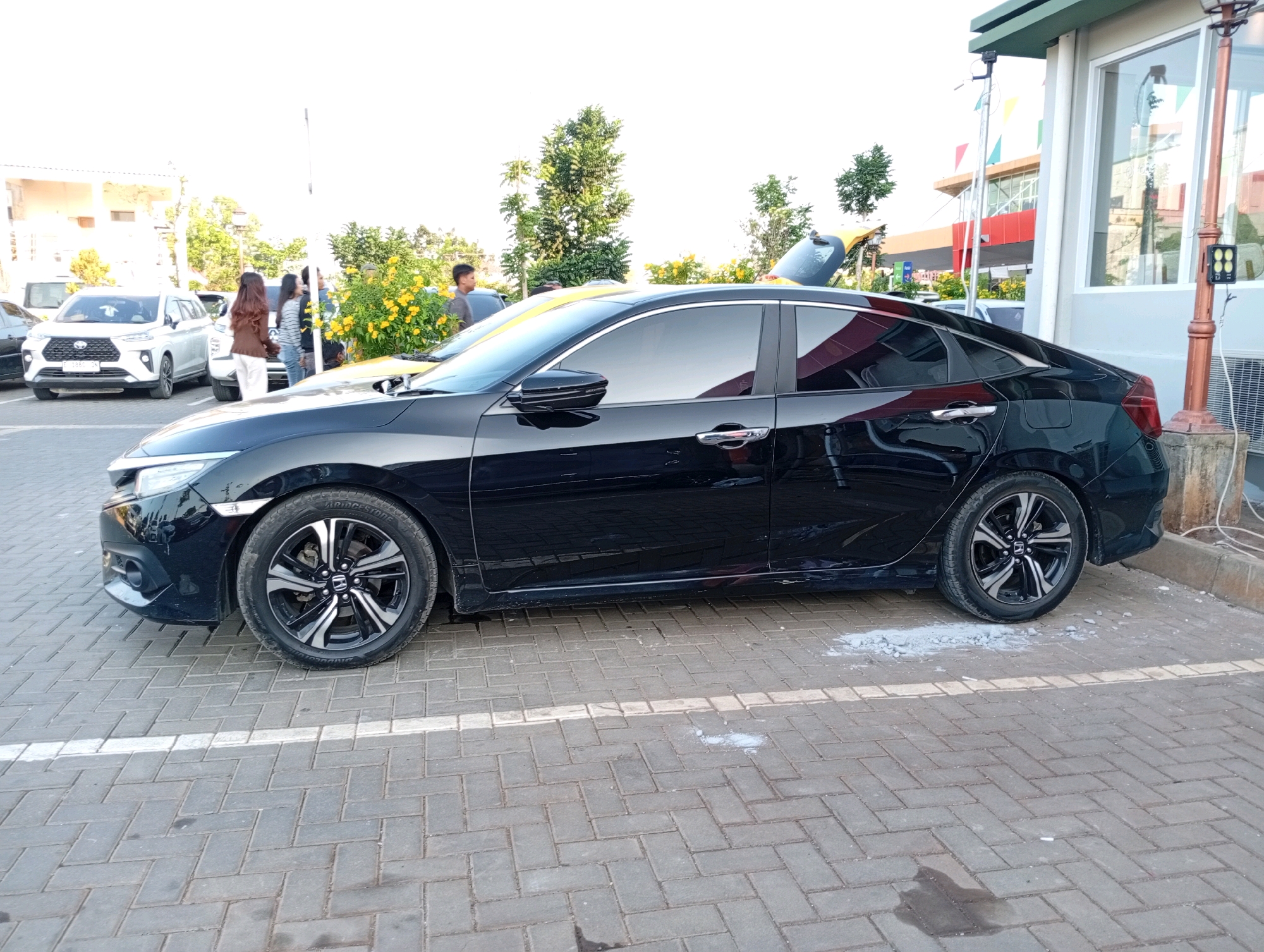 A black sedan is parked on a brick-patterned surface, in what appears to be an outdoor parking area. The car is positioned to the right, angled towards the viewer. Its front left wheel and part of the front bumper are visible. The car's windows are tinted. Several other vehicles, including a white SUV, are parked in the background. Two women and a man are standing near the white SUV, seemingly engaged in conversation. Lush green trees and yellow flowering bushes are visible behind the vehicles. In the far background, a modern building with a prominent red section and large glass windows can be seen. The scene suggests an outdoor commercial or public space.
