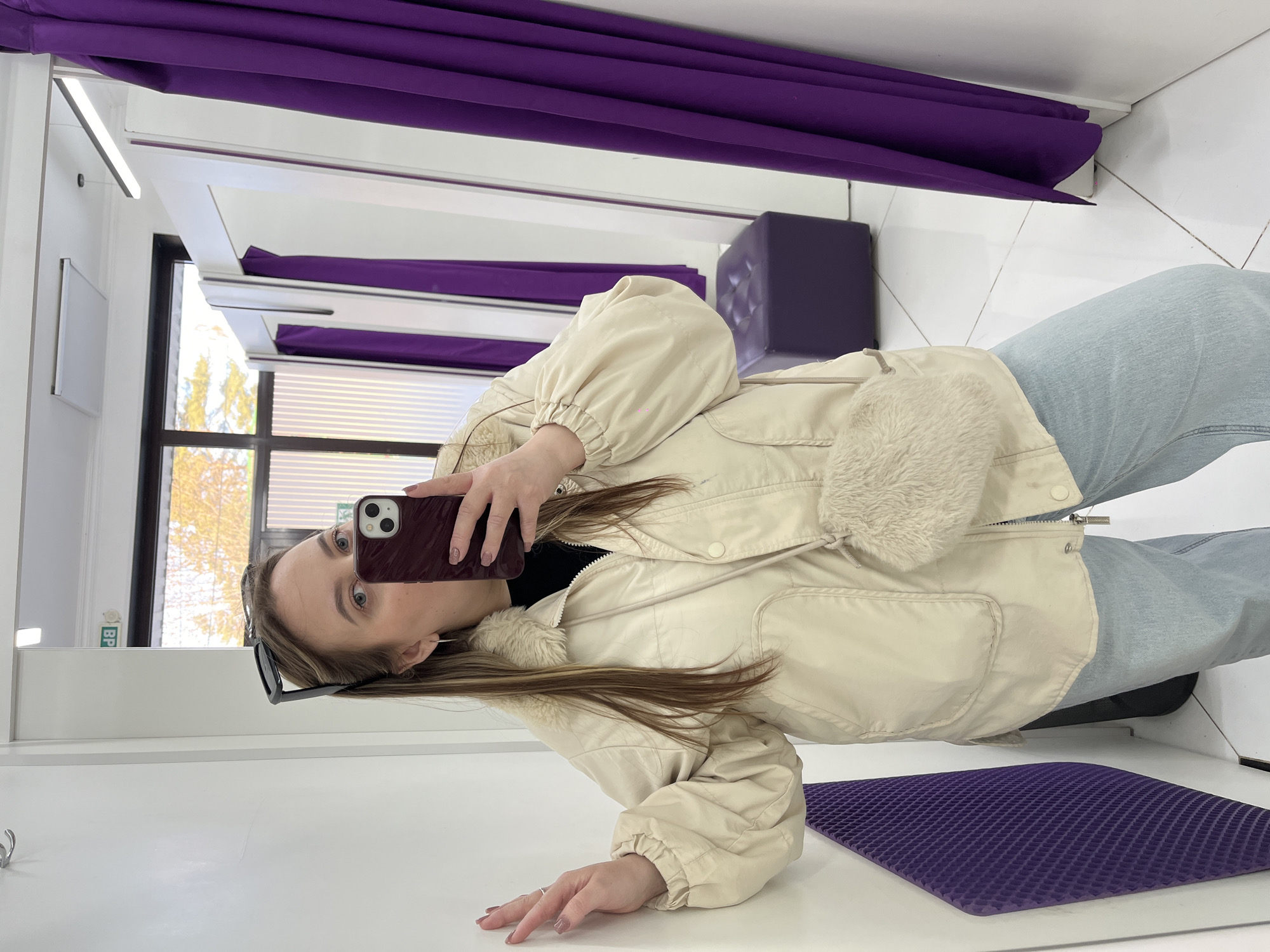 The image shows a woman taking a selfie in what appears to be a dressing room. She is holding a burgundy-colored smartphone, and her face is visible in the reflection. She's wearing sunglasses on her head, a cream-colored jacket with fur trim, a black top, and light-wash jeans. Behind her, there's a purple curtain, a purple ottoman, and a window showing trees outside. There's also a purple seat cushion. The location may be Novopokrovskaya, Russia, based on additional context.