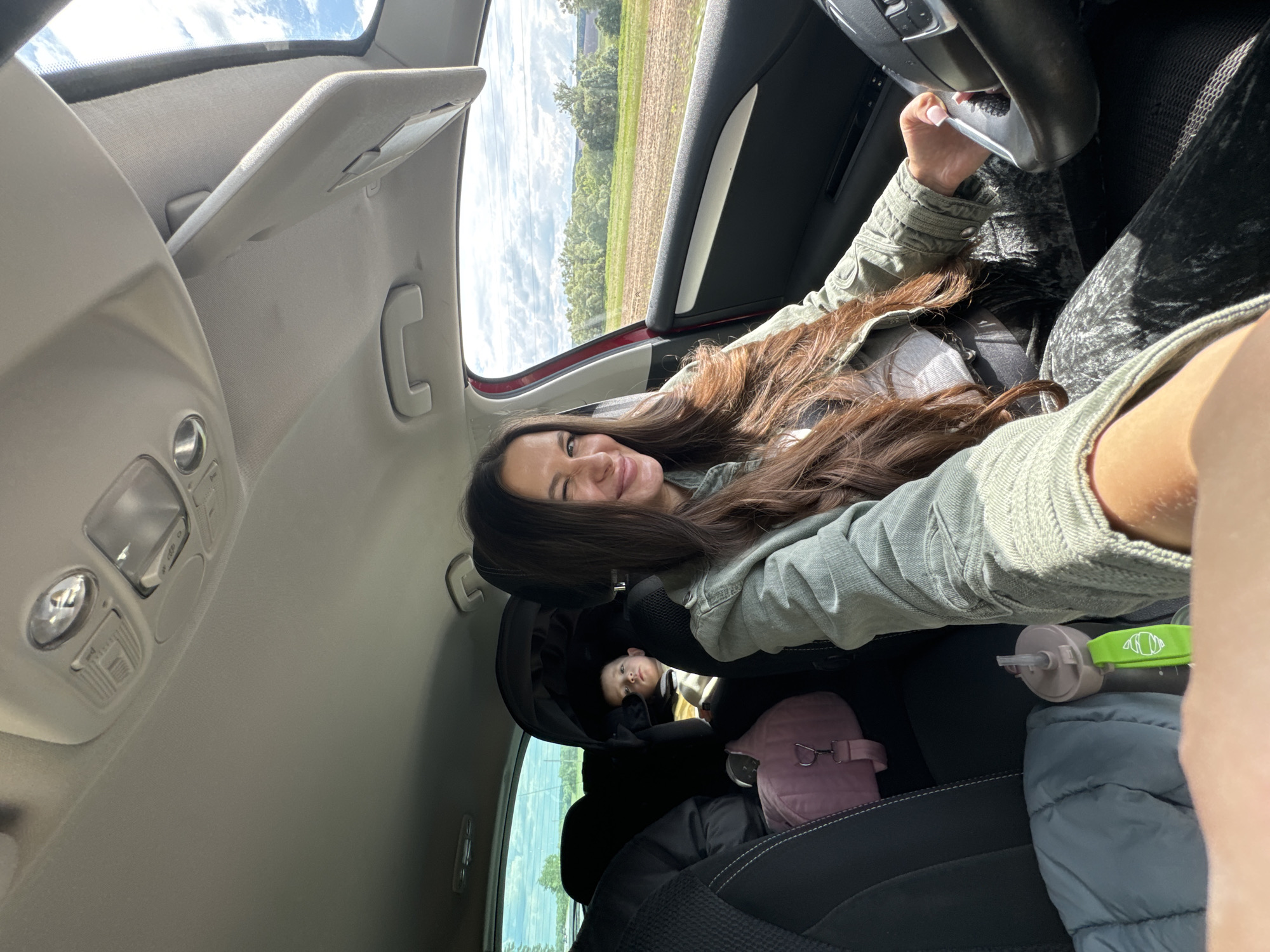 A woman sits in the driver's seat of a car, turned towards the camera, winking and smiling. She has long brown hair, wearing a light green jacket over a grey shirt. In the backseat is a baby in a car seat, a pink purse, a water bottle, and a grey puffy jacket. The landscape visible through the car windows suggests a rural area. The car's interior lights and sun visor are visible. The location is Kremyonki, Russia.
