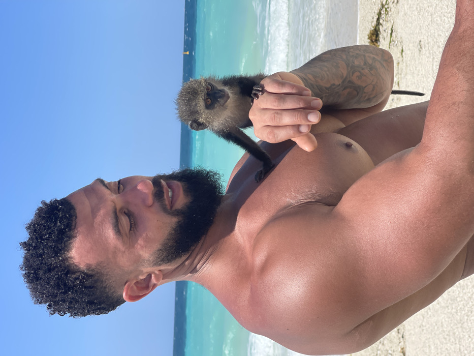 A medium-close shot depicts an adult male holding a small vervet monkey on a sunny beach in Zanzibar, Tanzania. The man, positioned on the left side of the frame, has dark, curly hair with a faded cut, a full beard, and a muscular build with tanned skin. A tattoo is visible on his left forearm. He is shirtless, facing slightly to his right, with his head tilted downwards, mouth slightly open, and eyes partially closed, appearing to engage with the monkey. The monkey, greyish-brown with a dark face, stands perched on the man's outstretched left hand, with its left arm extended towards his chest. Its gaze is directed slightly downwards and to the right. The background features a vibrant turquoise ocean with small waves breaking on a white sand beach. A single yellow buoy is visible in the distant water. The sky is clear and blue.