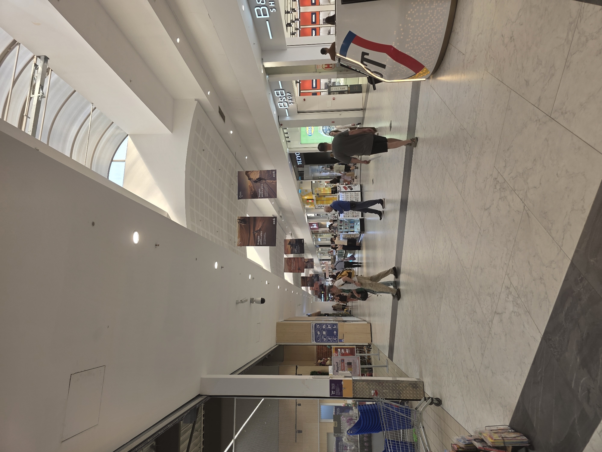 This is a photograph of the interior of a modern shopping mall, likely in Bucharest, Romania. The mall features a bright, open-air design with a large, arched skylight allowing natural light to flood the space. The flooring is a light-colored marble tile, and the walls are painted white. Various shops and businesses are visible along the sides of the mall, indicated by signage such as "B&B SHOP" and "TEZYO." Several people are walking through the mall, some singly and some in pairs. One man is walking away from the camera, wearing a grey t-shirt and shorts. Another man, wearing a khaki shirt and pants, walks towards the camera with a backpack. A shopping cart is partially visible on the left side of the image. Advertising banners hang from the ceiling, featuring landscape imagery. The overall impression is one of a clean, well-lit, and busy commercial environment.