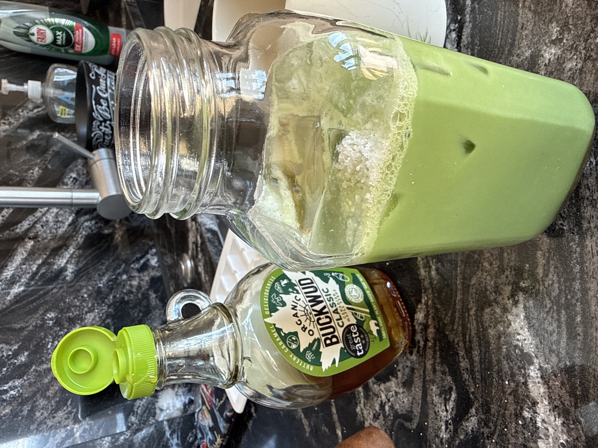 The scene depicts a domestic kitchen countertop, situated in London, United Kingdom. The primary surface is a dark, veined countertop, indicative of granite or quartz. Two main vessels are positioned prominently: a clear glass bottle of "ORGANIC BUCKWUD CLASSIC" maple syrup, approximately half-filled with an amber liquid, featuring an open, bright green flip-top cap; and to its right, a clear, square-bottomed glass jar containing a vibrant green, ice-filled beverage, likely matcha, topped with white granular material and foam. Visible in the background are a metallic sink faucet, a clear pump dispenser bottle, a black bowl with partial white text ("Quest... Be Queen"), and a green and white bottle of "FAIRY MAX" dish soap. The presence of "FAIRY MAX" dish soap, a common household product in the UK, and the "Great Taste 2023" award label on the maple syrup, a UK-based food award scheme, are consistent with a location in the United Kingdom.